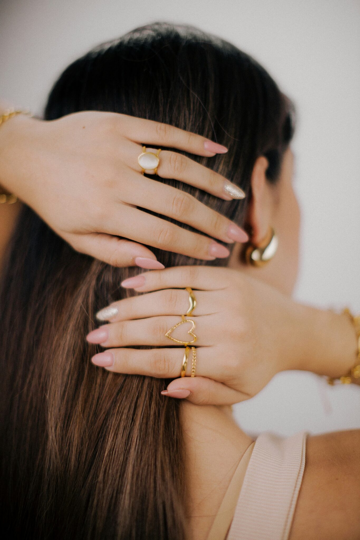 A person with long brown hair holds the back of their head, displaying manicured nails and several gold rings and bracelets.