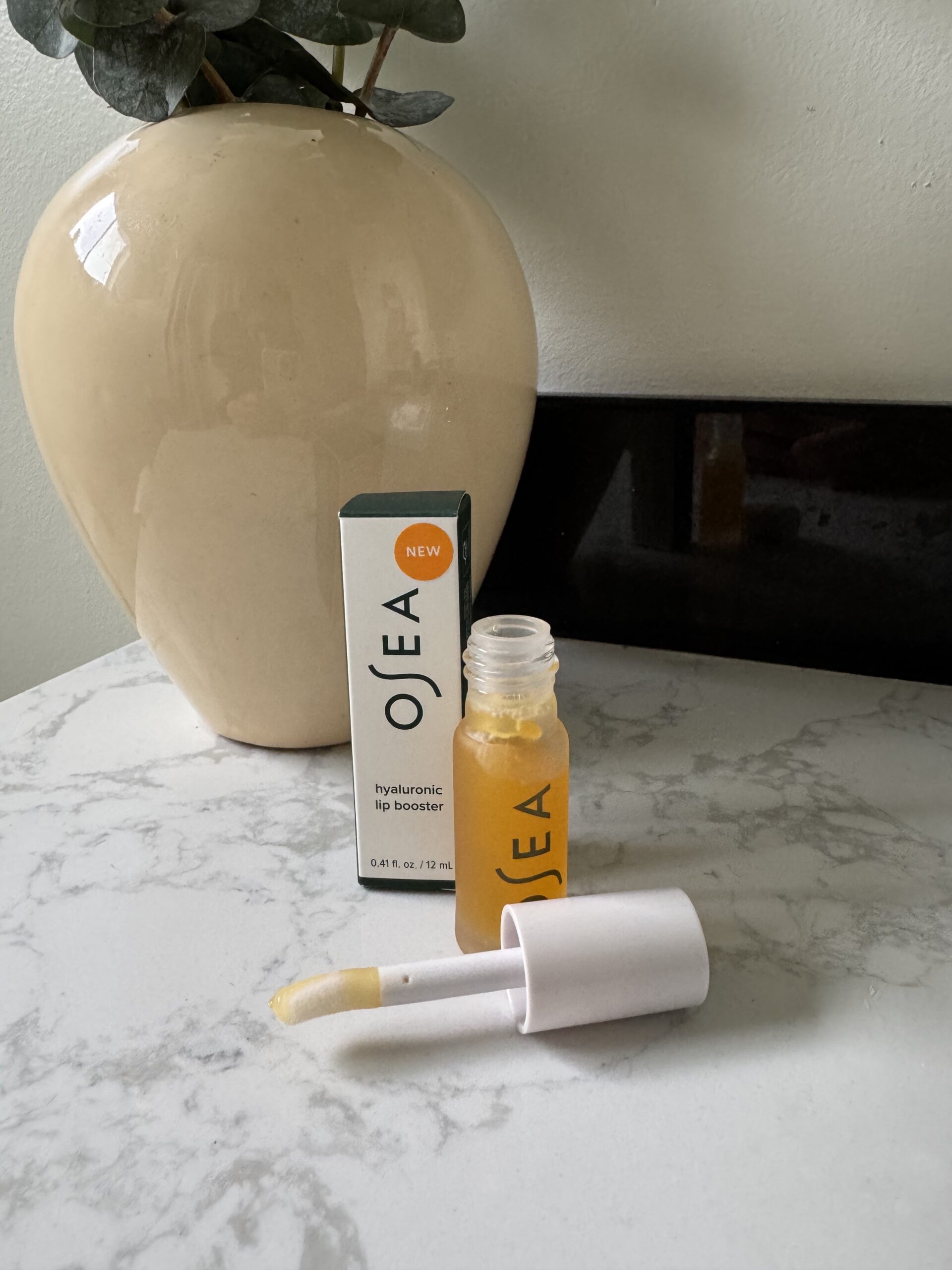 A bottle of OSEA hyaluronic lip booster with its applicator wand lying in front, next to the product box on a marble surface with a ceramic vase in the background.