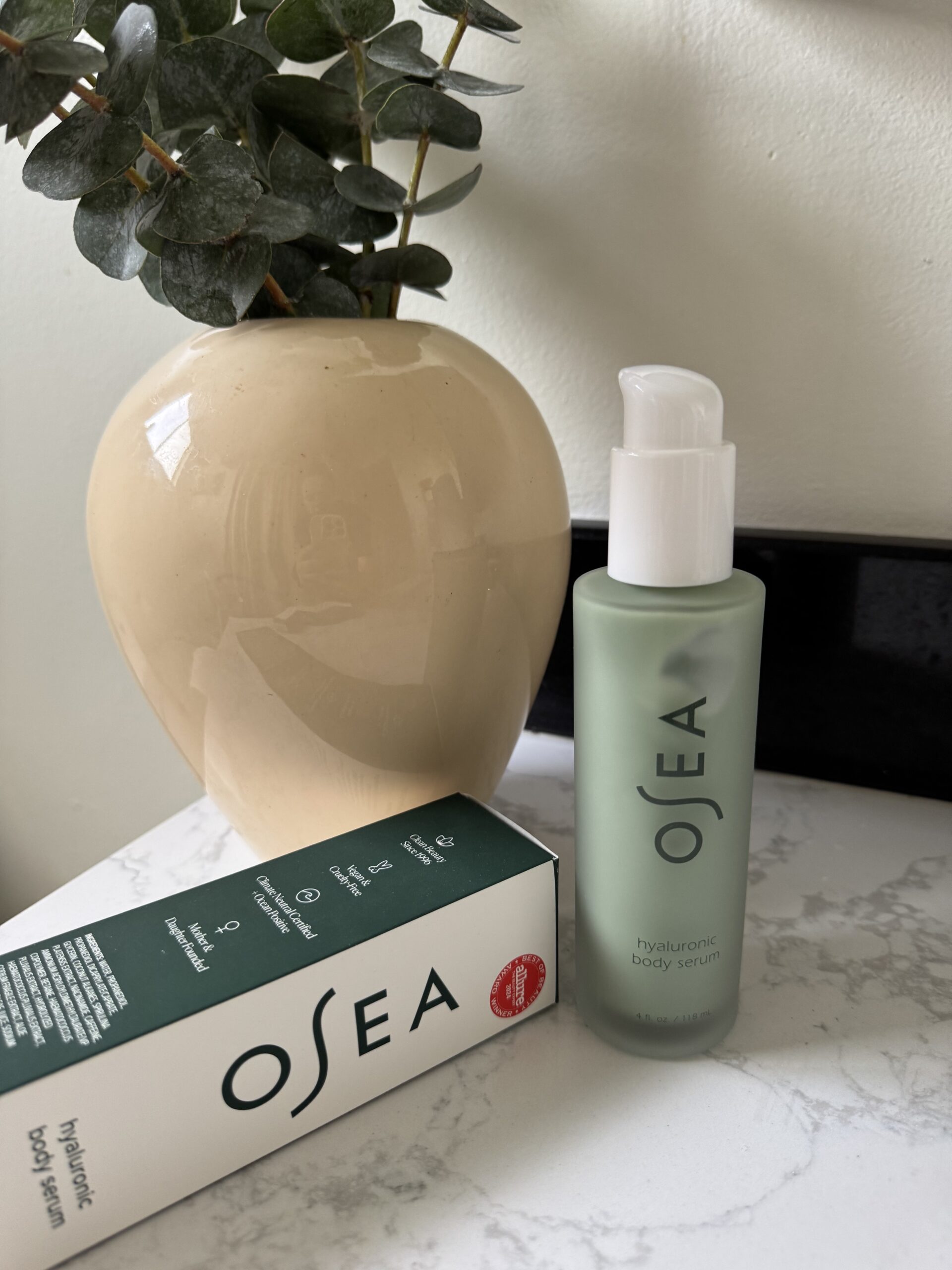 A bottle of OSEA hyaluronic body serum stands next to its box on a marble surface, with a beige vase and green plant in the background.