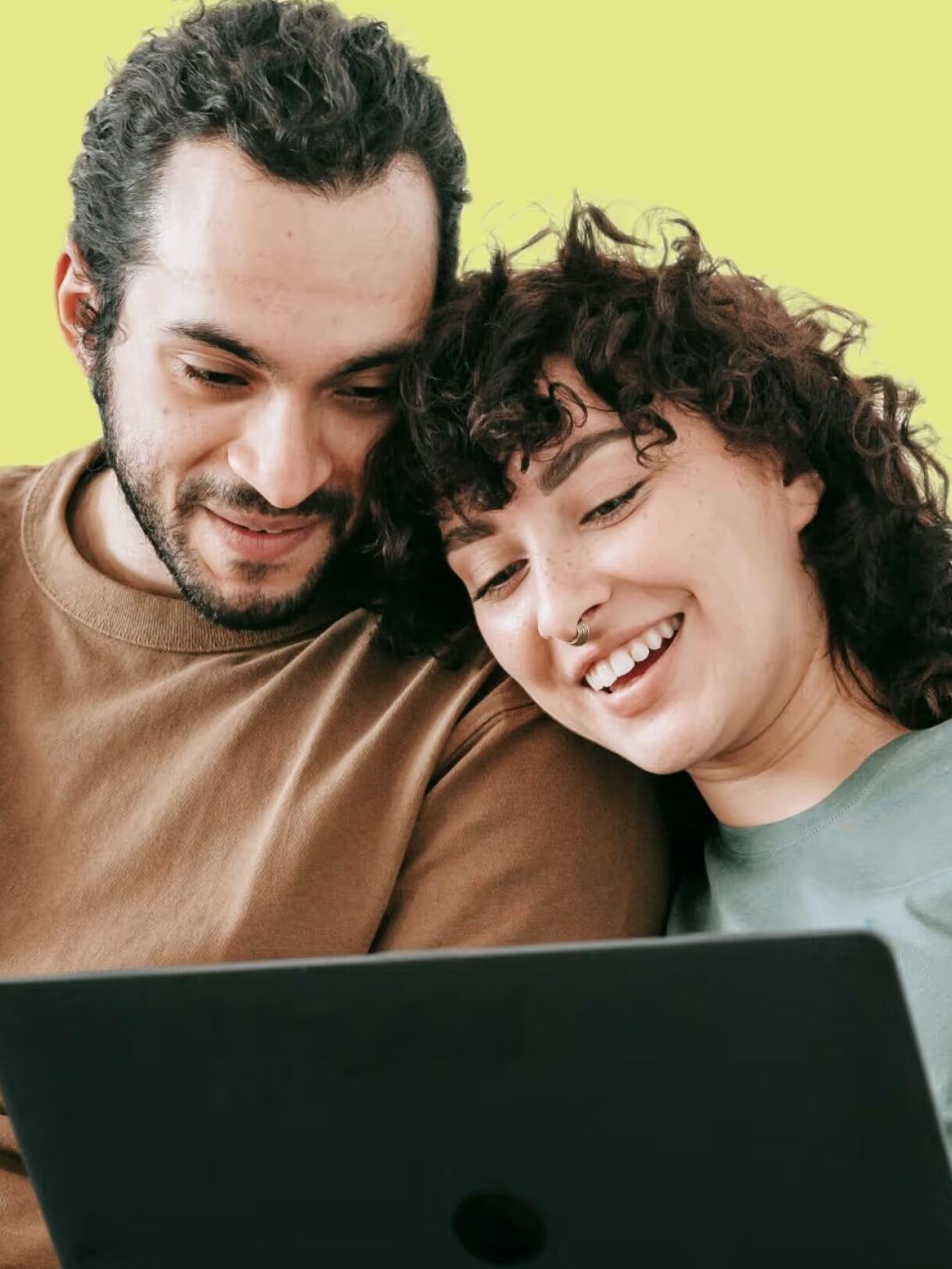 A man and a woman sit closely on a sofa, smiling while looking at a laptop. The background is a solid light green.