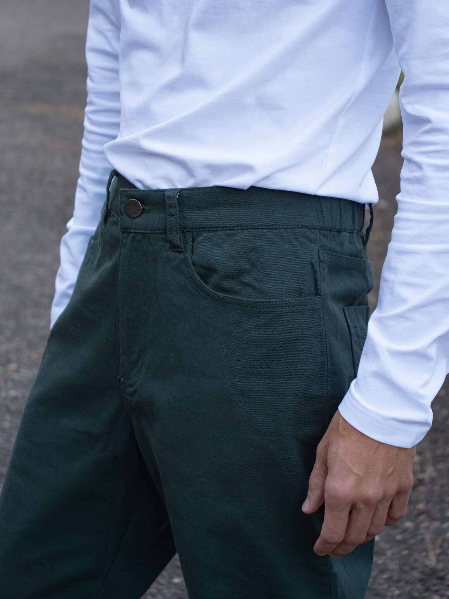 A person wearing a white long-sleeve shirt and dark green pants stands outdoors with one arm relaxed by their side.