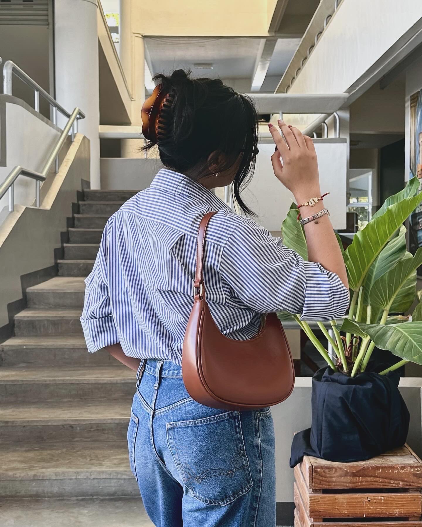 Woman with a brown shoulder bag and striped shirt stands near stairs, facing away, holding up her hand. There is a large potted plant beside her.