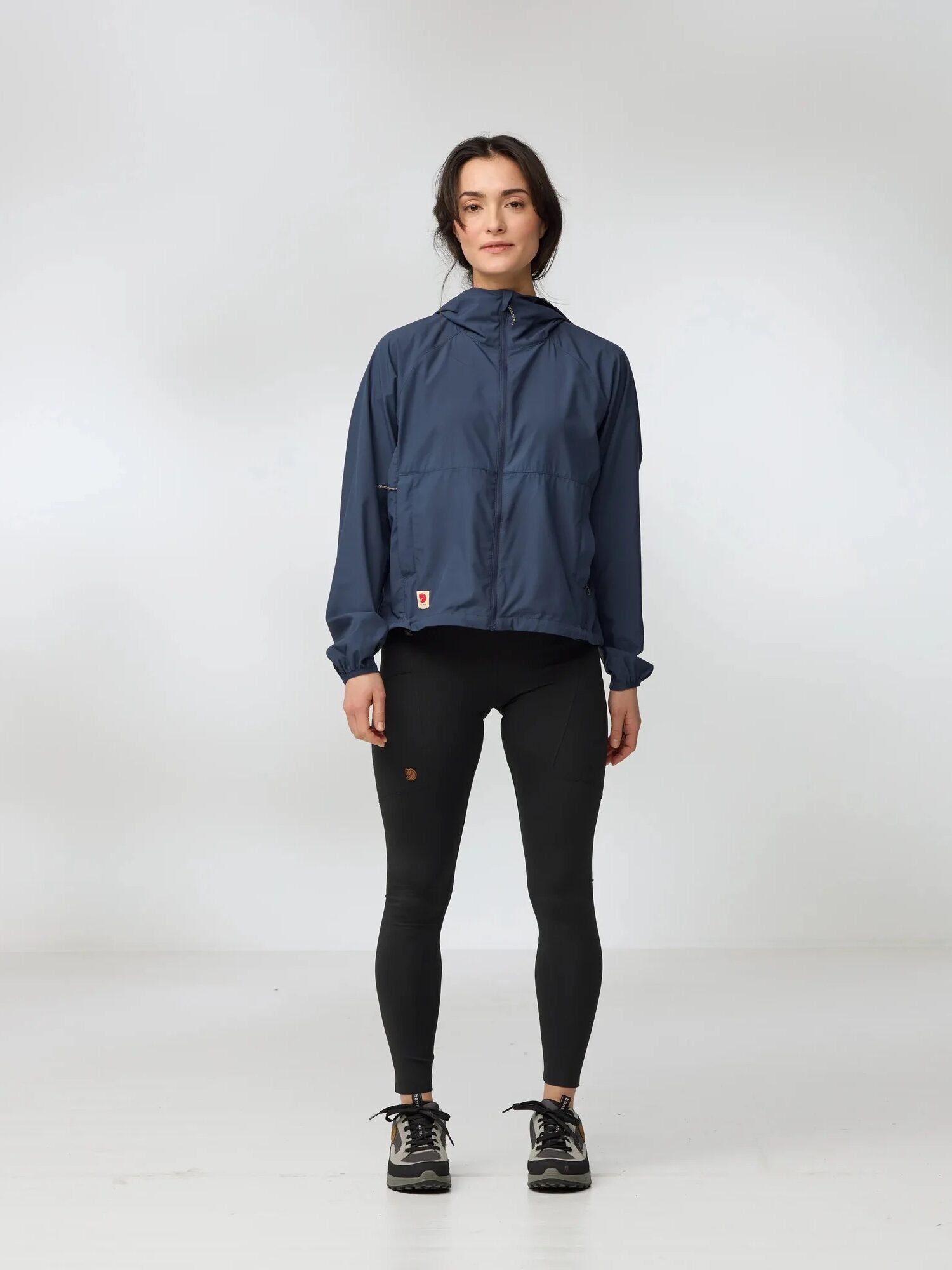 A woman stands indoors against a plain white background, wearing a navy waterproof raincoat, black leggings, and black sneakers.