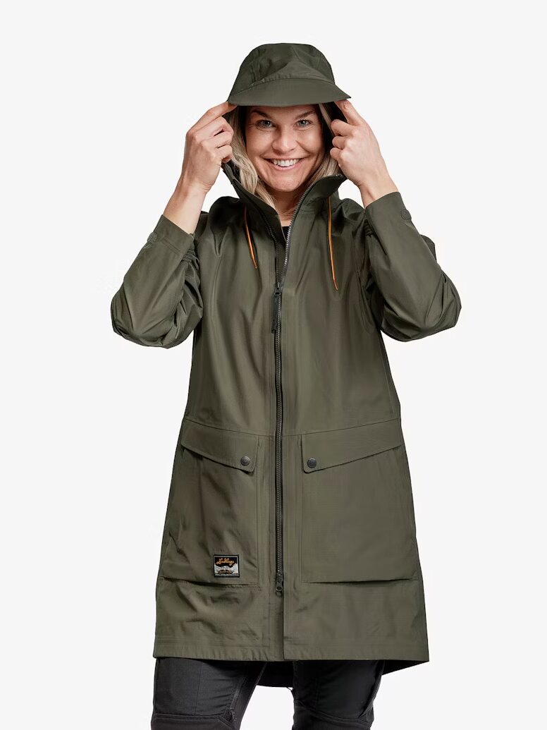 A person wearing an olive green waterproof raincoat and matching hat, smiling and holding the sides of the hood, stands against a plain white background.