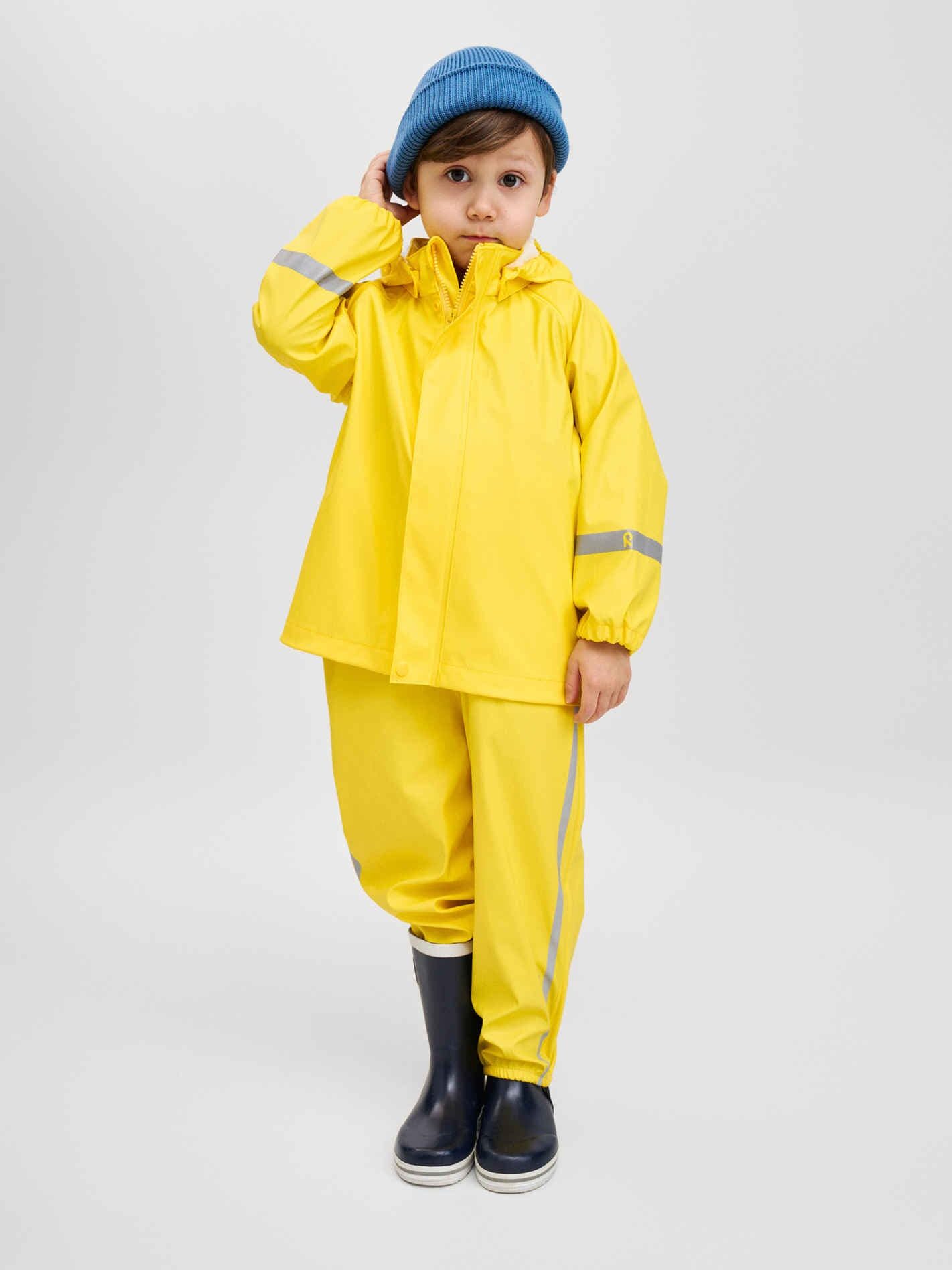 A young child wearing a waterproof yellow raincoat and pants, blue beanie, and black rain boots stands against a plain light background.