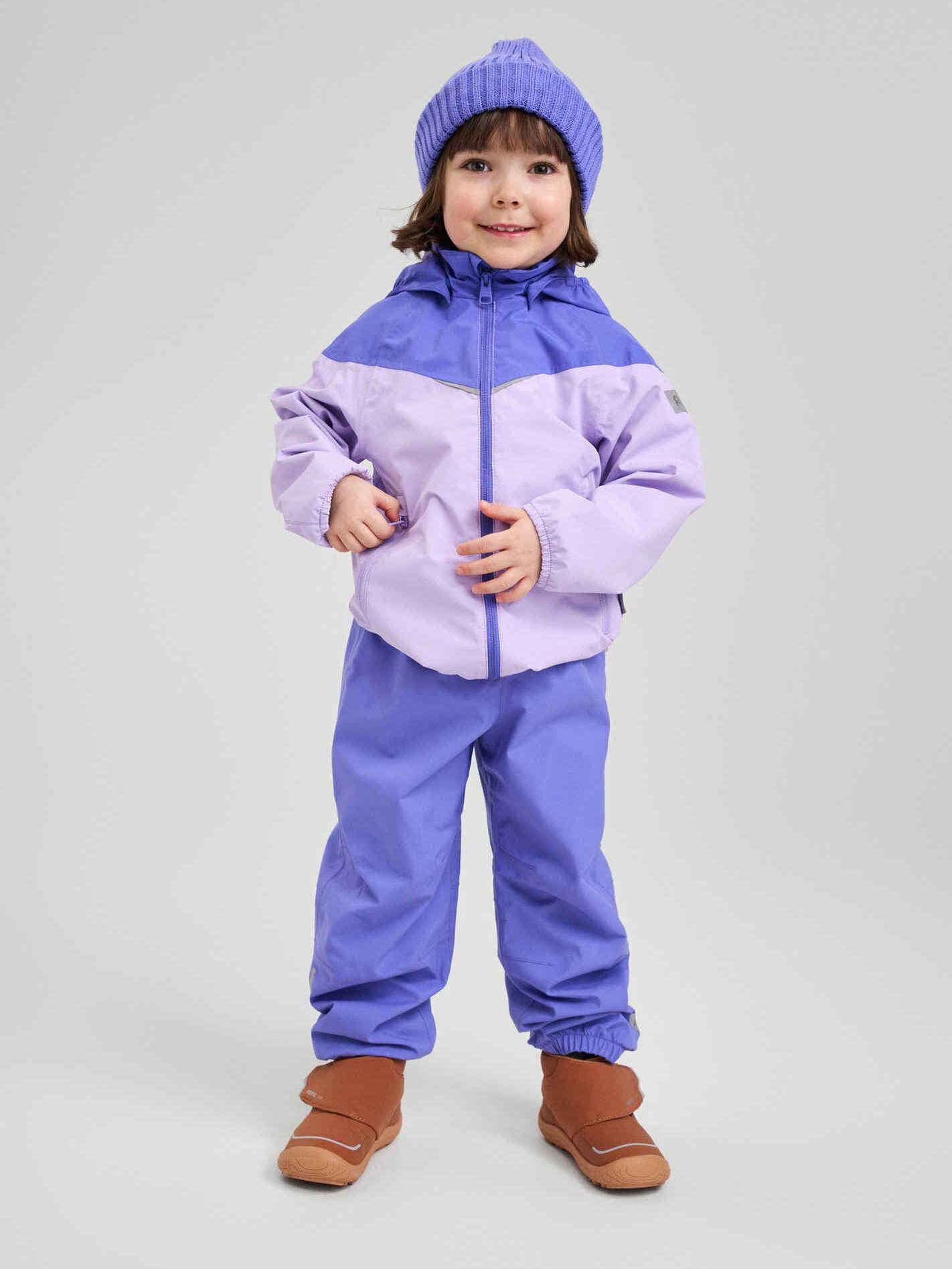 Young child standing and smiling, wearing a purple beanie, a light purple waterproof raincoat and lavender jacket, matching pants, and brown boots against a plain gray background.