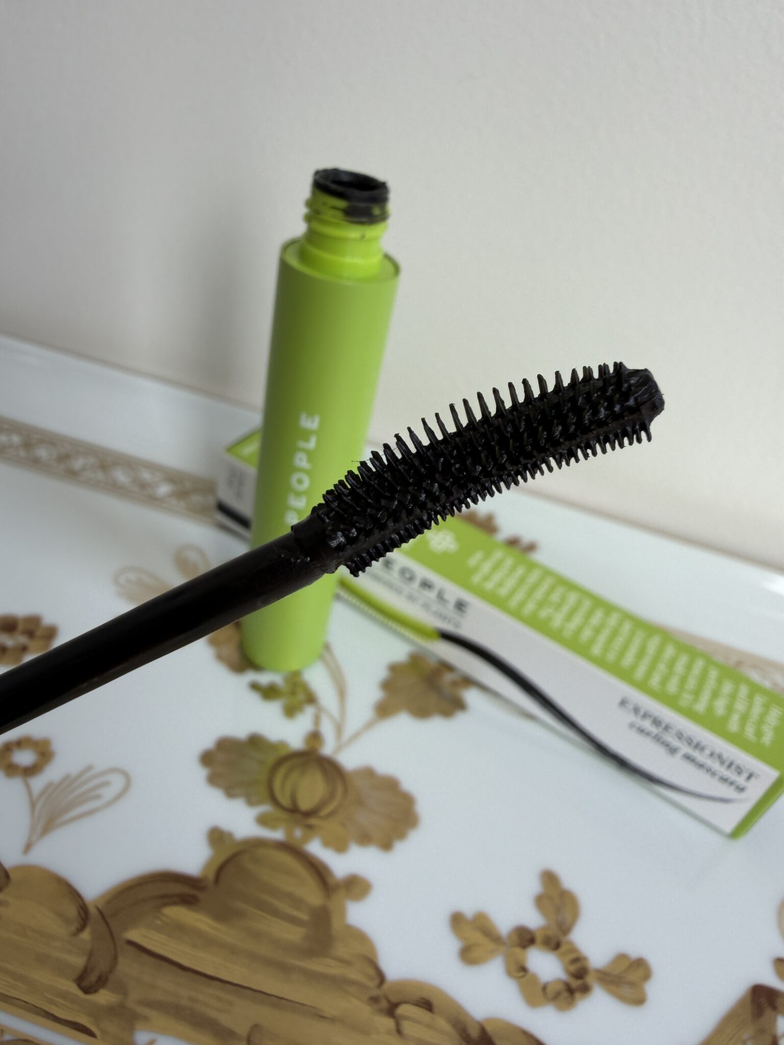 A close-up of a black mascara wand with bristles, held above an open green mascara tube, placed on a decorative tray with a floral design.