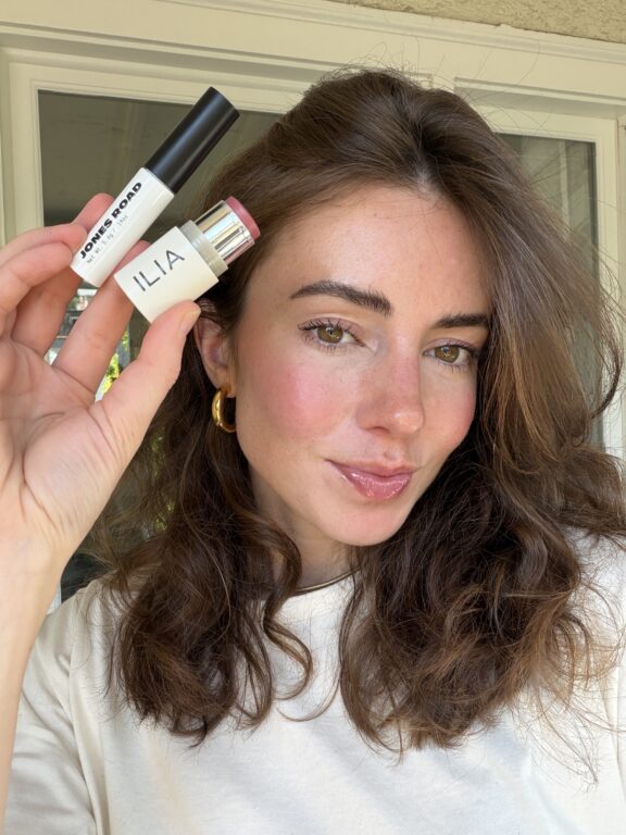 A woman with wavy brown hair holds up a Jones Road mascara and an ILIA blush stick, showing her makeup look with rosy cheeks and glossy lips.