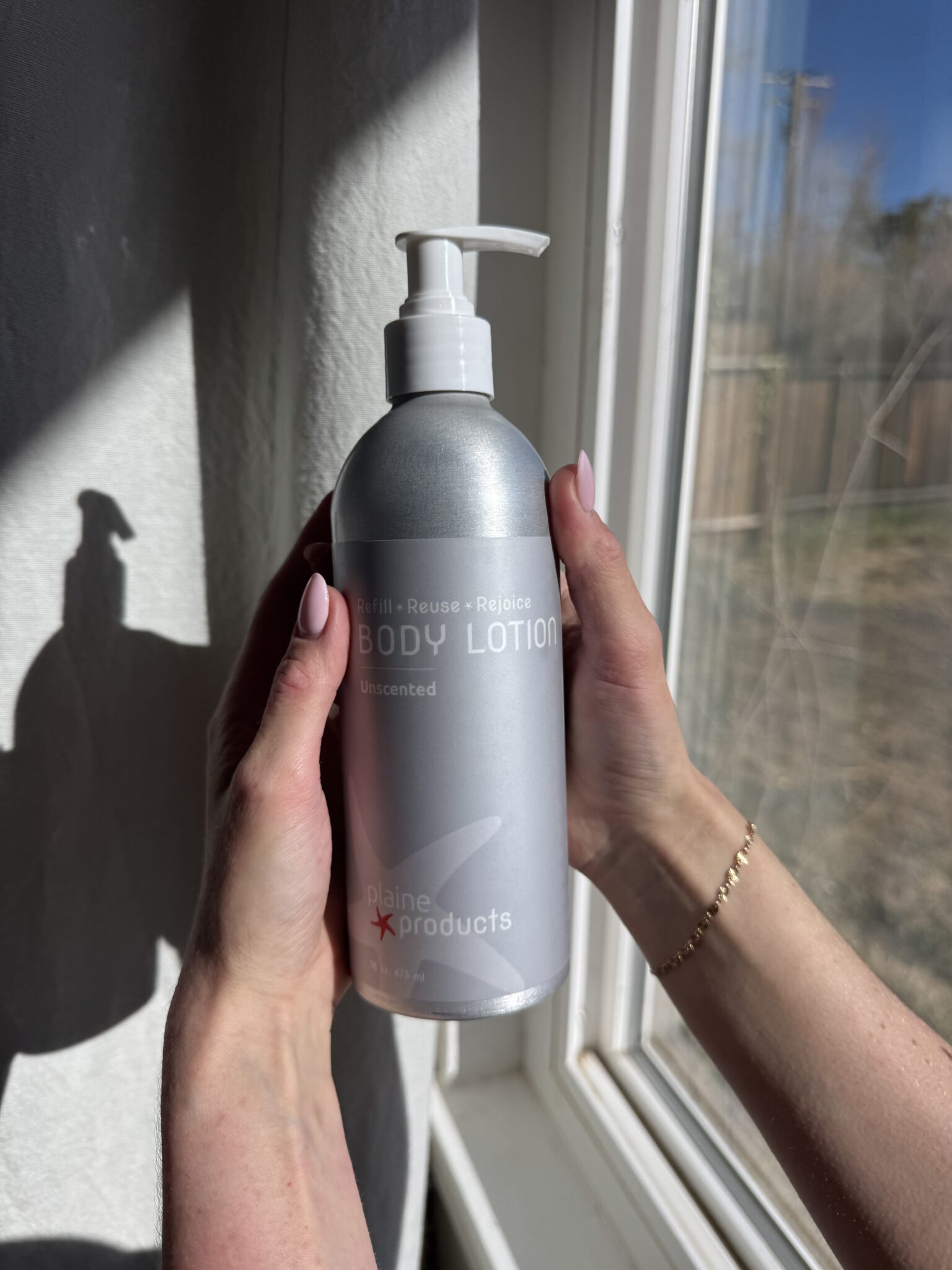 Two hands are holding a silver bottle of Plaine Products unscented body lotion near a sunlit window.