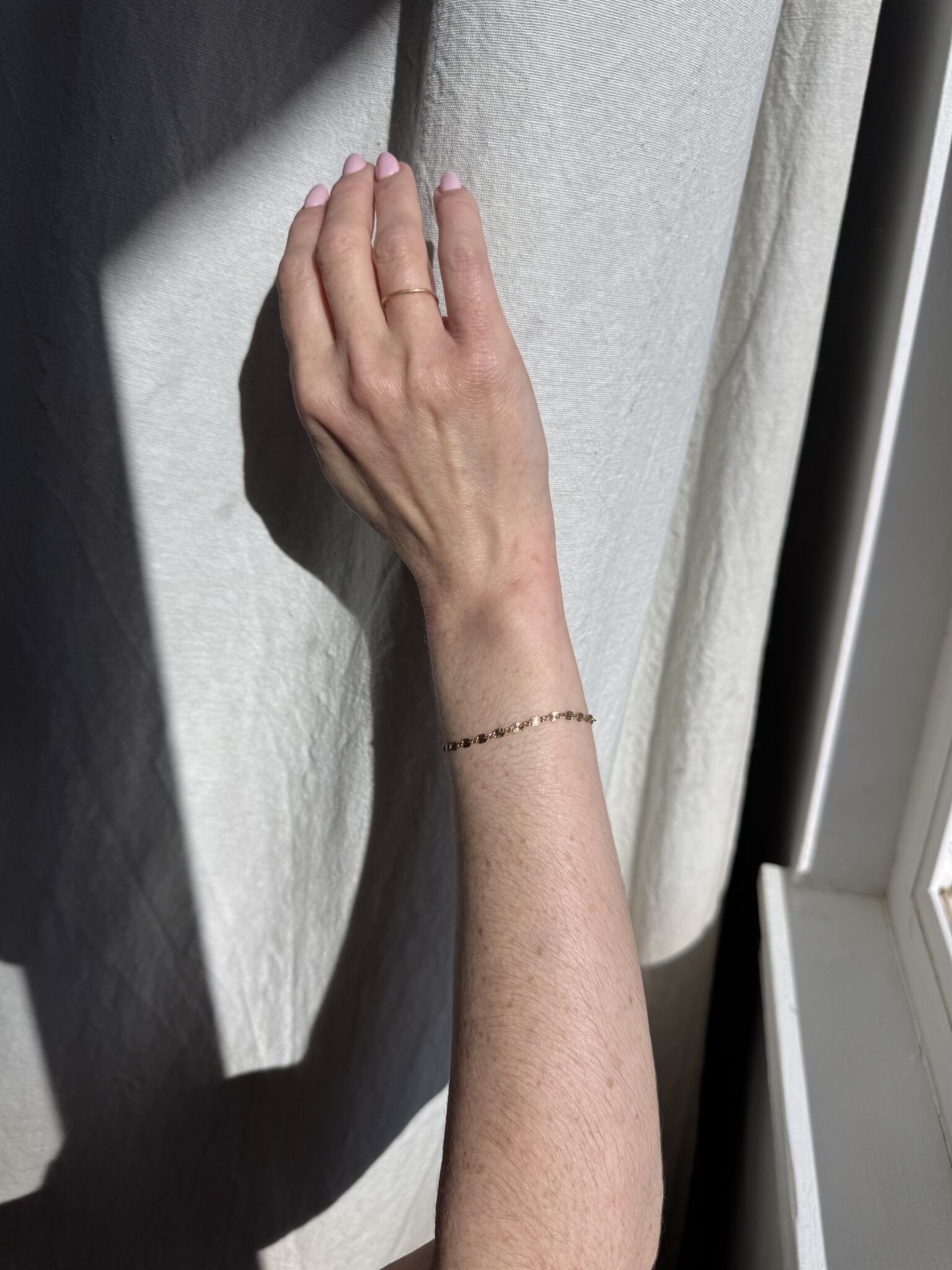 A hand with pale pink nails and a thin beaded bracelet is extended toward a light-colored curtain in sunlight.