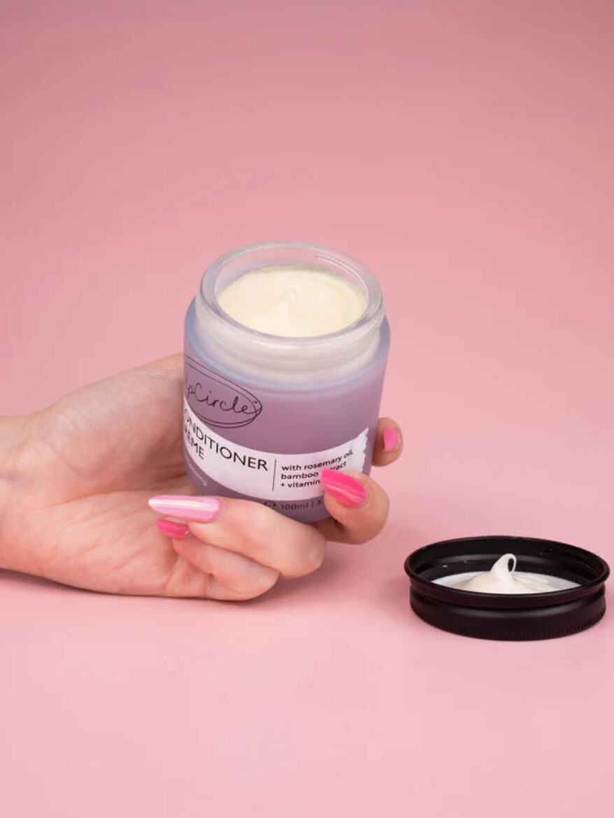 A and holding an open purple jar of hair conditioner in front of a pink background. By Plastic Freedom.