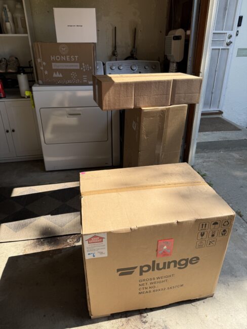 Several cardboard boxes of various sizes, including ones labeled "plunge" and "Honest," are stacked in a laundry area near a washer and dryer.