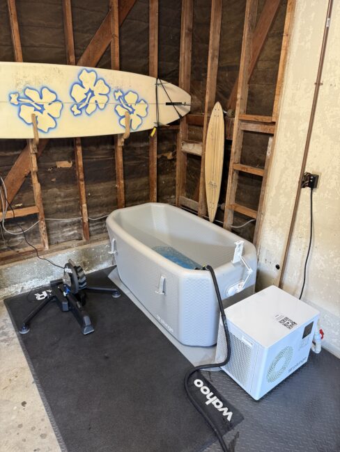 A gray inflatable ice bath filled with water sits in a garage next to a cooling unit, with two surfboards mounted on the wall behind it and a bike trainer on the left.