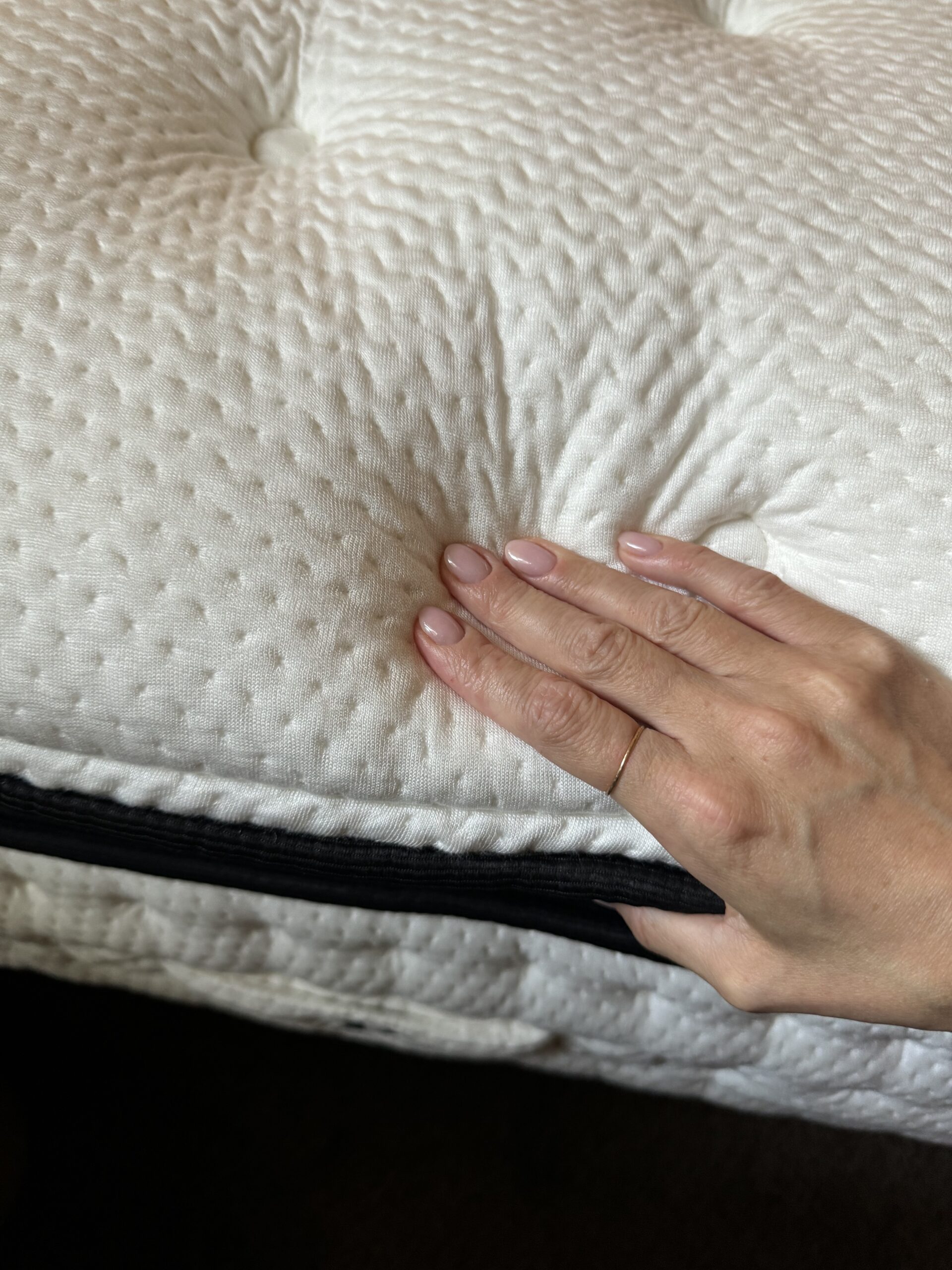 Kate Arceo's hand presses down on a nontoxic Plushbeds mattress with a black zipper edge, reflecting its commitment to safety and comfort.
