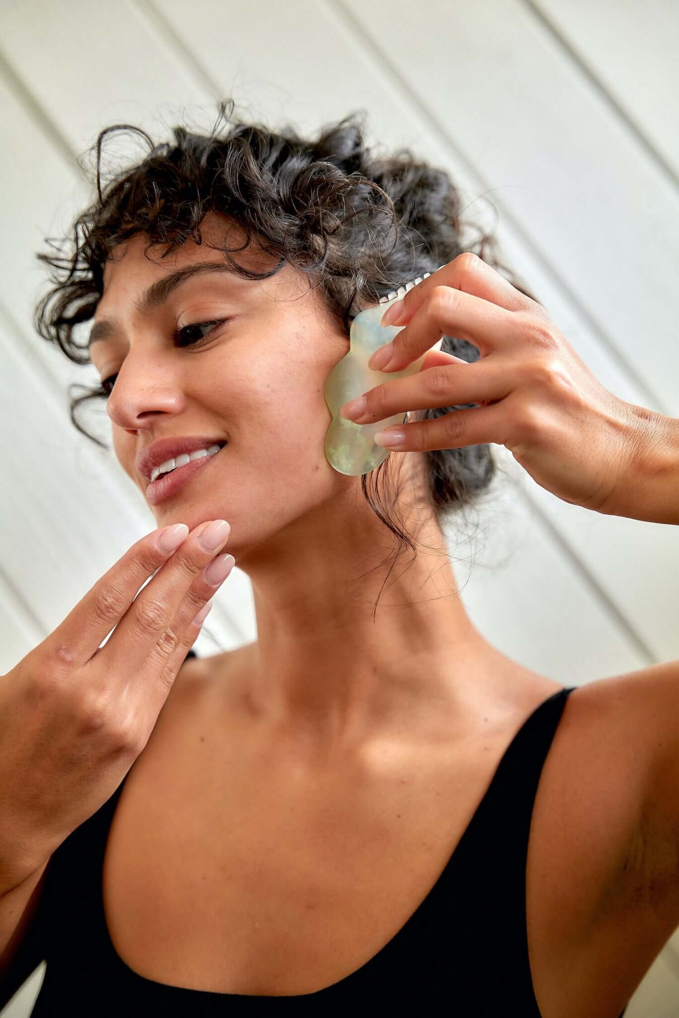 A woman uses a jade gua sha tool to massage her face, holding it against her cheek near her ear, with a slight smile.