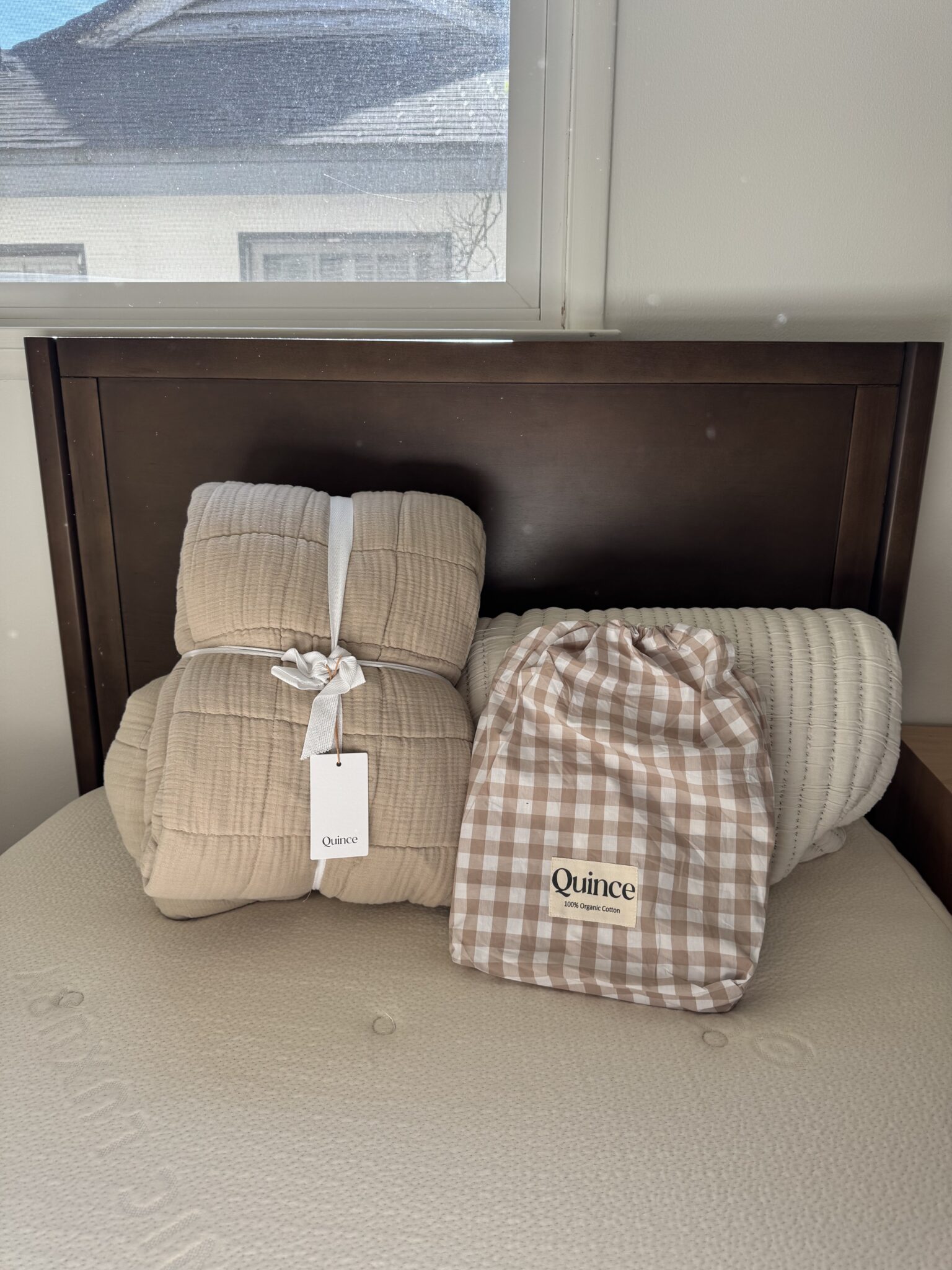 A beige quilt, a white bolster pillow, and a gingham Quince bag are placed on a mattress against a dark wood headboard under a window.