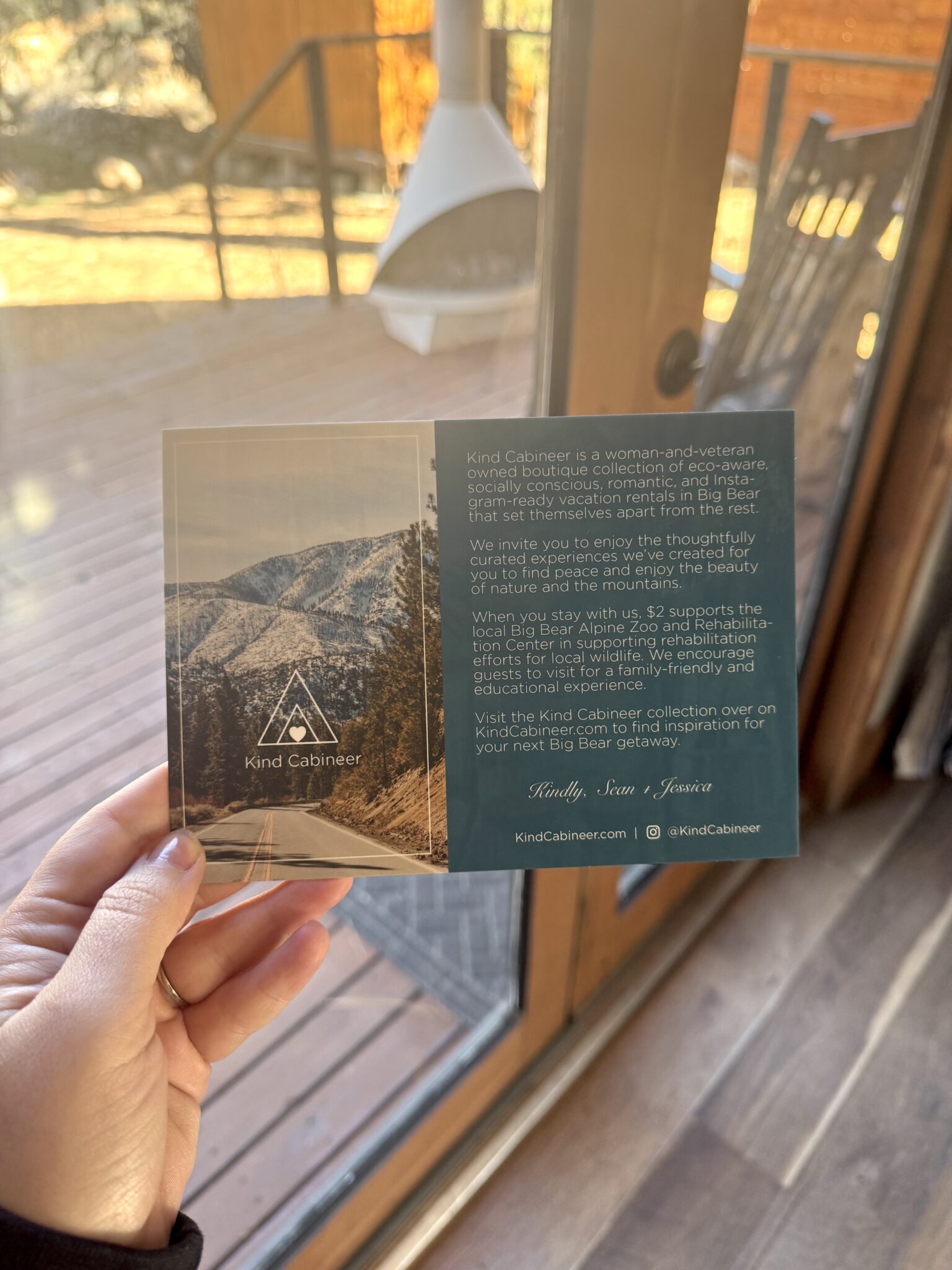 A hand holds a Kind Cabineter postcard by a glass door; the card features a mountain photo, logo, and text describing vacation rentals.