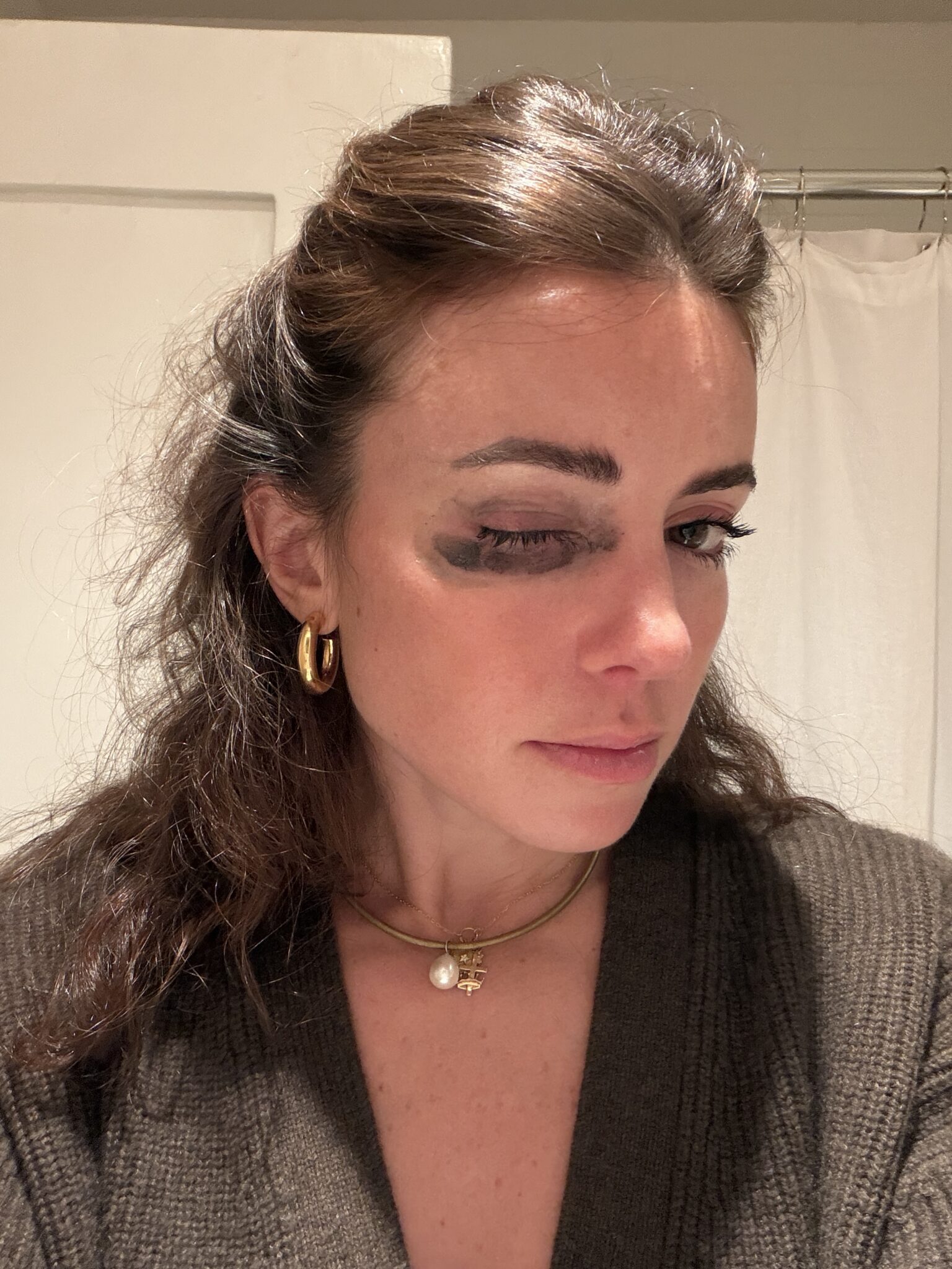 A woman with brown hair and gold jewelry has smeared black makeup under her right eye, standing indoors against a white background.