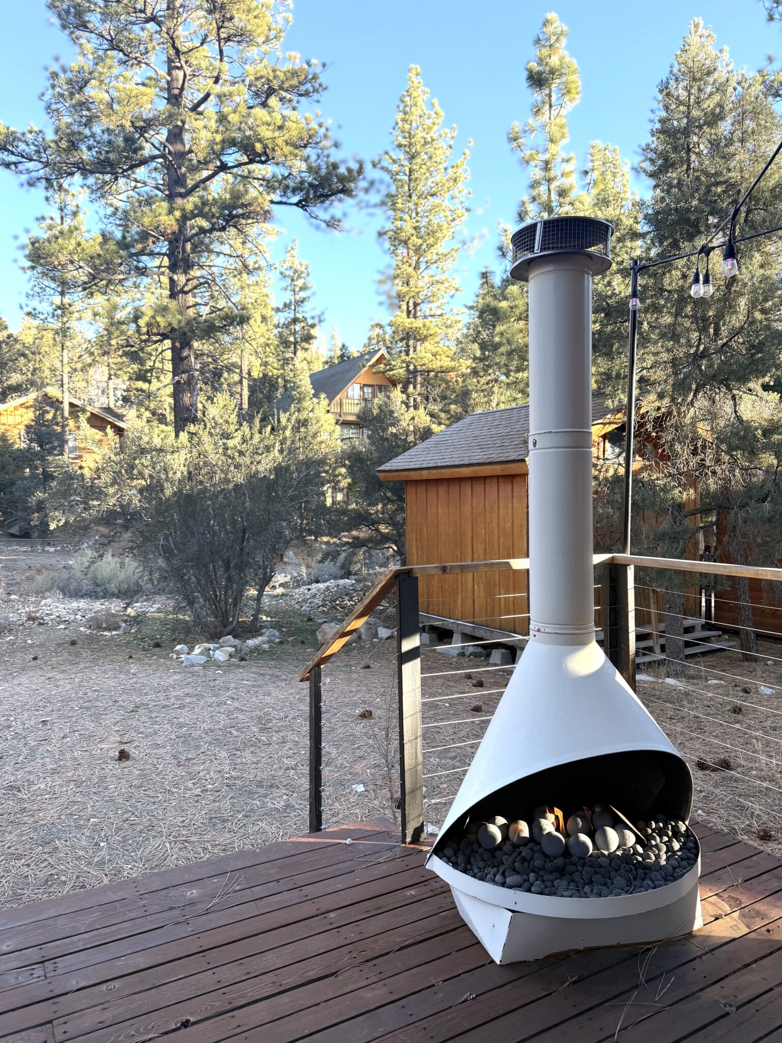 A white outdoor fireplace with a tall chimney sits on a wooden deck, surrounded by trees and cabin-style buildings in a forested area.