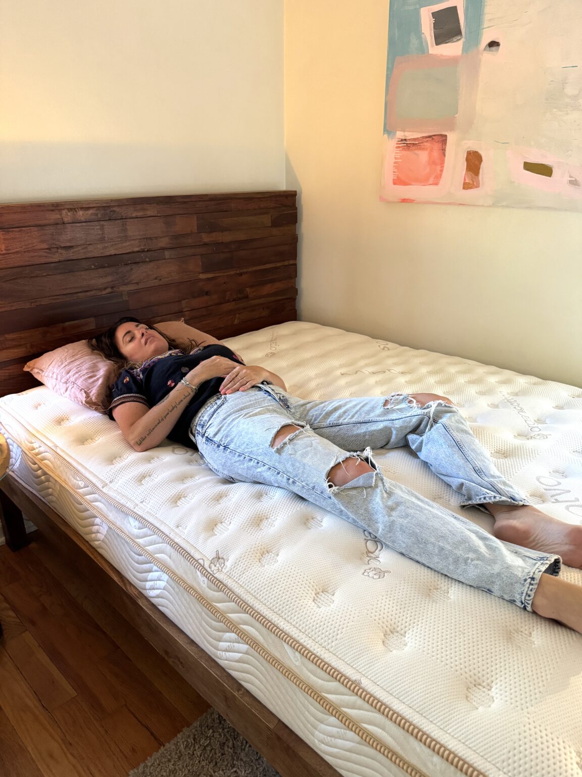 A woman in a black shirt and ripped jeans lies on her back on a bed with an organic mattress and a wooden headboard in a sunlit bedroom.