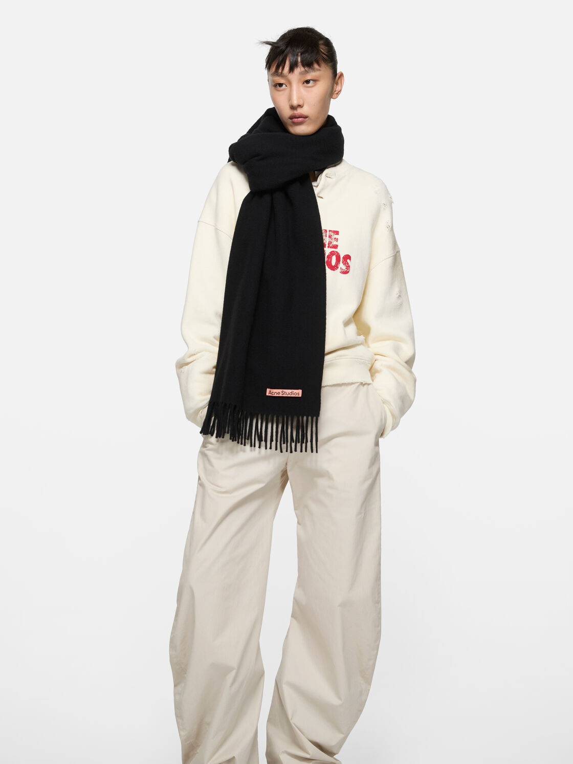 A person wearing a cream sweatshirt, wide-leg cream pants, black scarf, and black shoes stands against a plain white background.