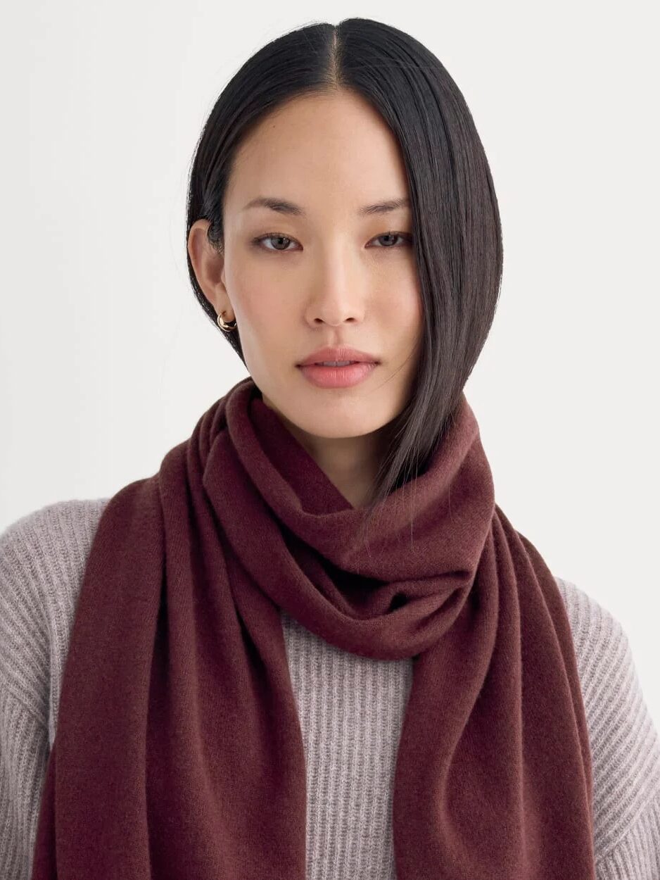 A woman wearing a taupe ribbed sweater and a dark brown scarf stands against a plain light background, looking directly at the camera.