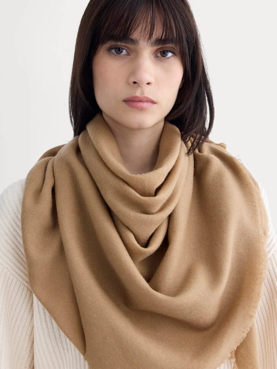 A person with straight dark hair wears a cream knit sweater and a large, camel-colored scarf draped around their neck, standing against a plain light background.