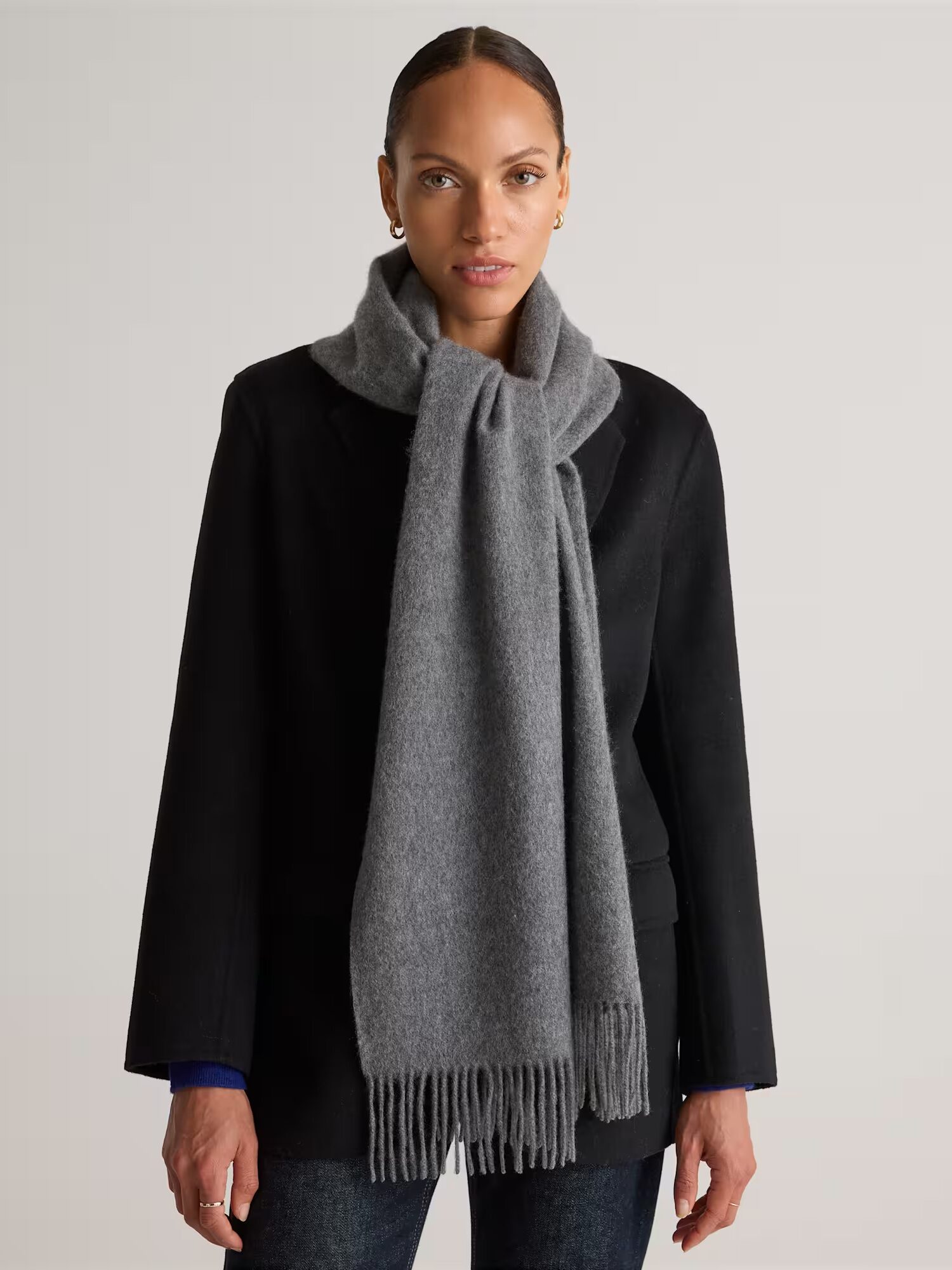 A person wearing a black coat and a thick gray scarf stands facing forward against a plain background.