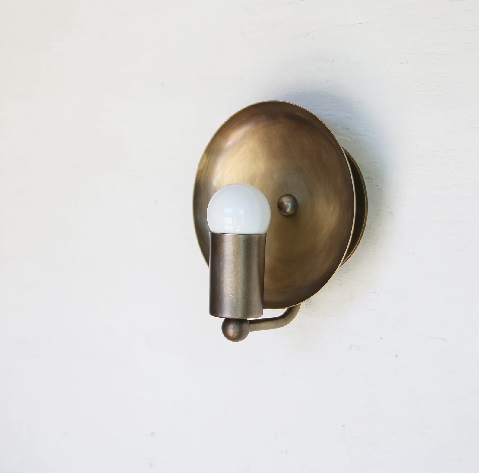 A wall-mounted brass sconce light fixture with an exposed round white bulb, attached to a round backplate on a white wall.