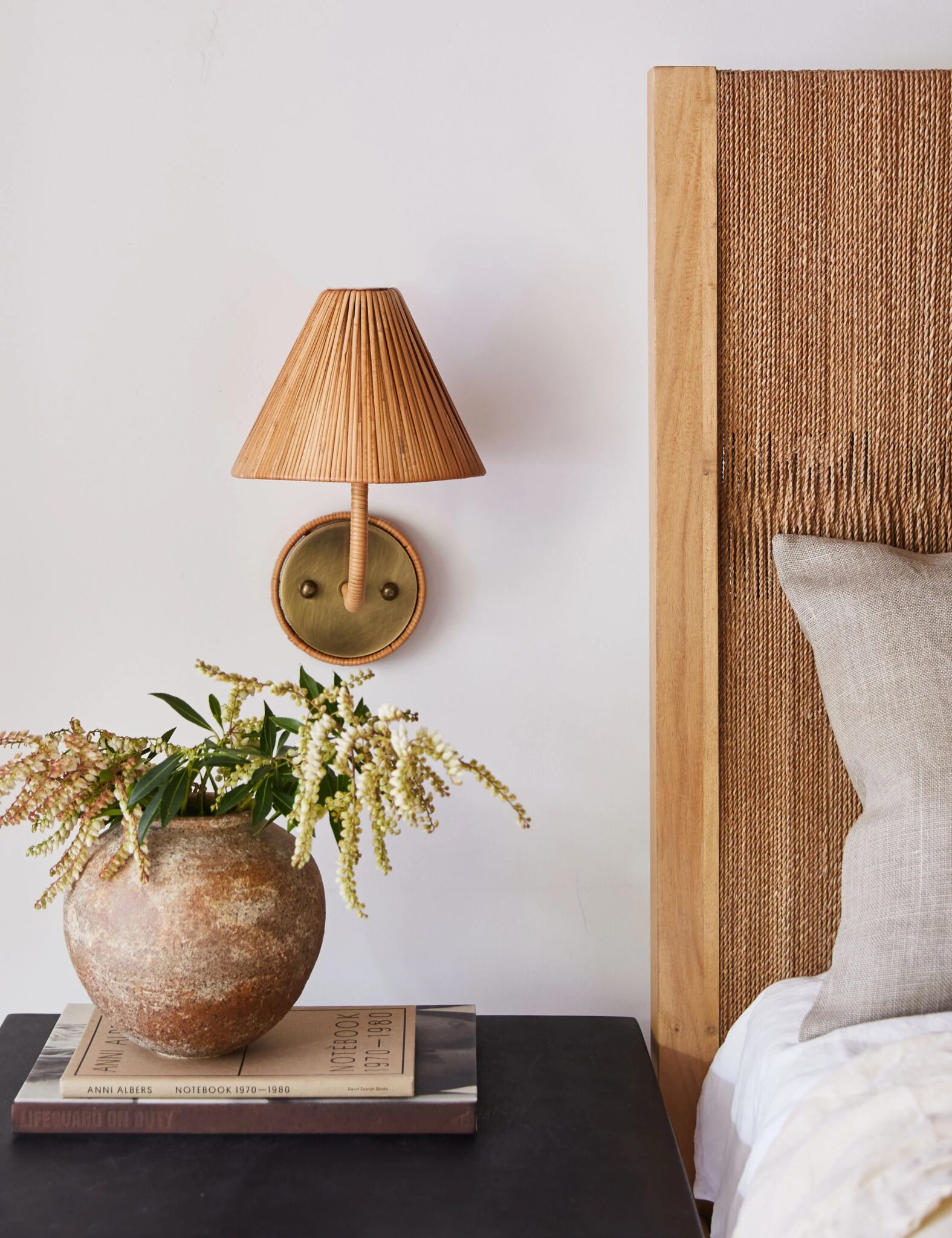 A bedside table with a rustic vase of flowers and books, next to a bed with a woven headboard and a wall-mounted wicker sconce lamp.