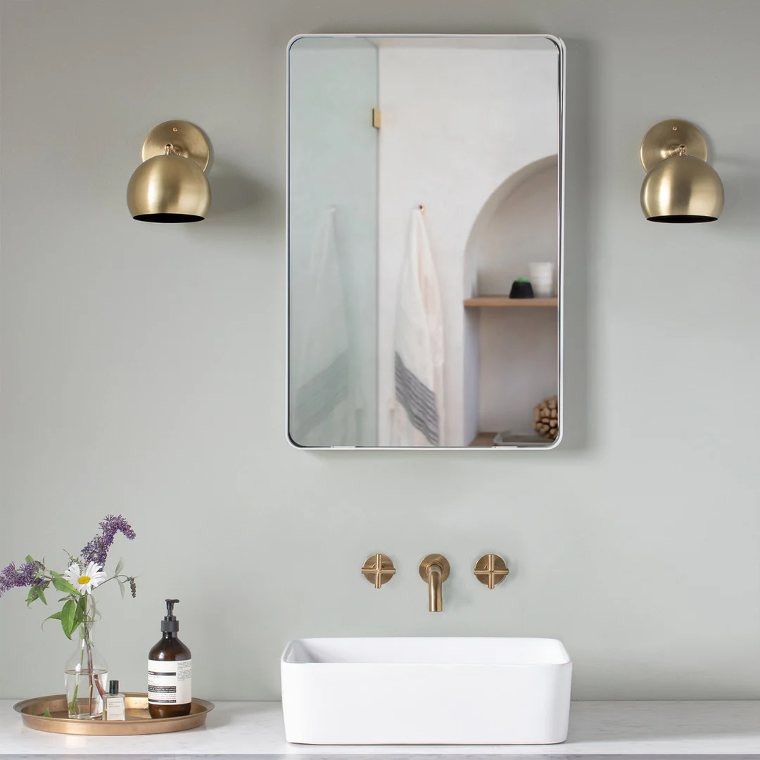 A modern bathroom with a rectangular mirror, gold wall sconces, a white basin sink, gold faucet, and a tray with a soap dispenser and vase of flowers.