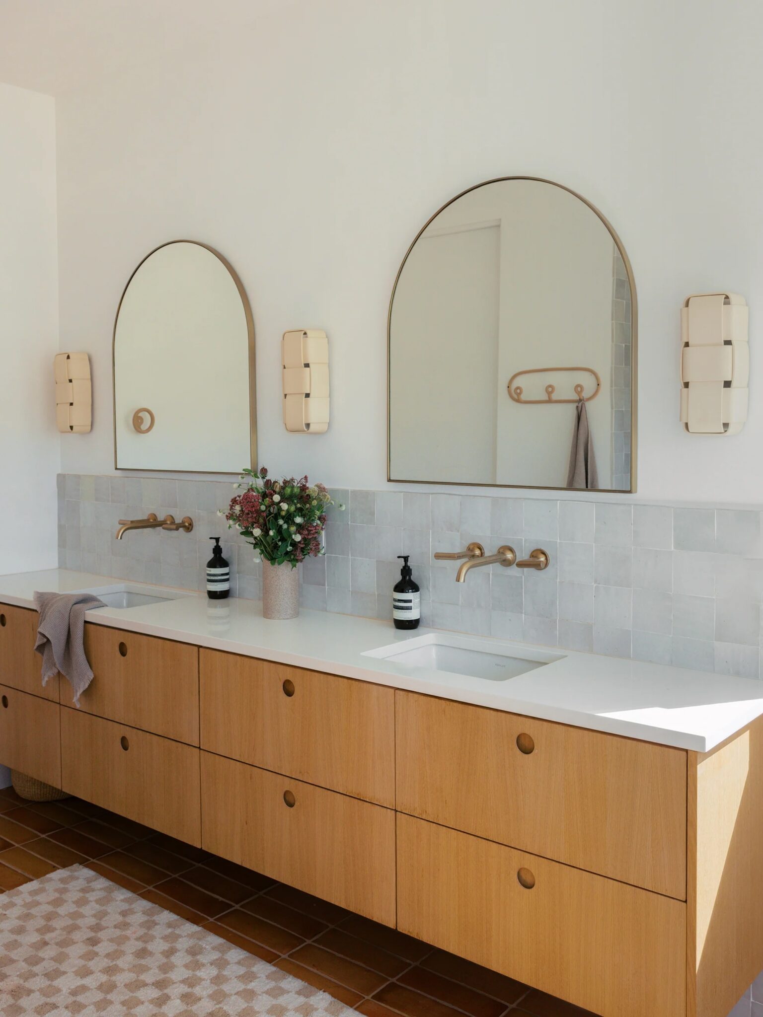 A modern bathroom with a double sink vanity, wooden cabinets, two large arched mirrors, wall-mounted faucets, soap dispensers, flowers, and tiled backsplash.
