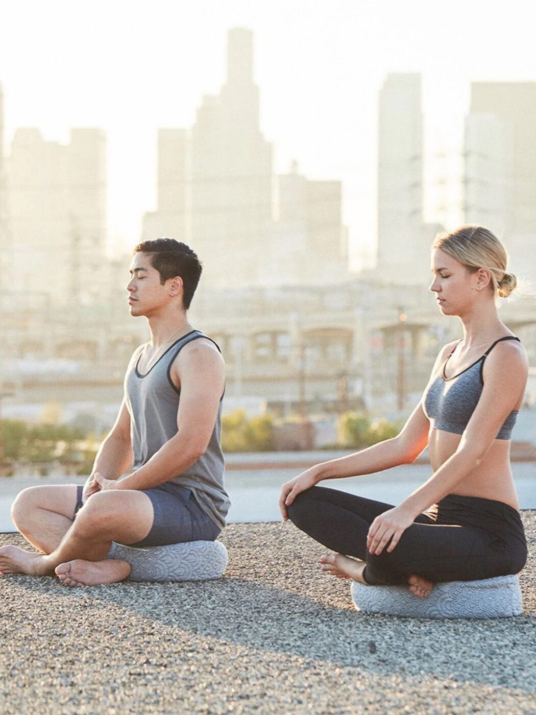 Two people meditating on mats outdoors, sitting cross-legged with a city skyline in the background.