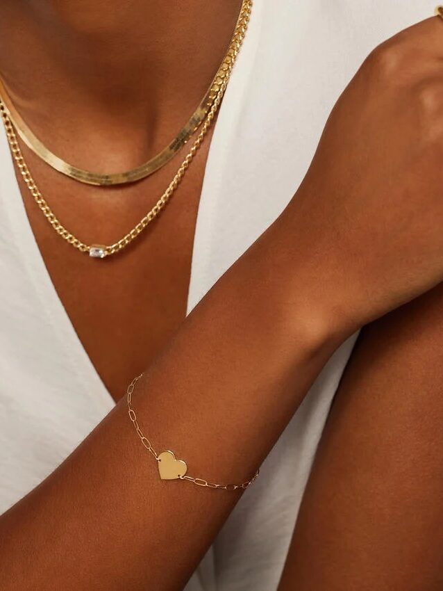 A person wearing a white top showcases gold jewelry: a chain necklace, a choker, a heart-shaped bracelet, and a simple gold ring.