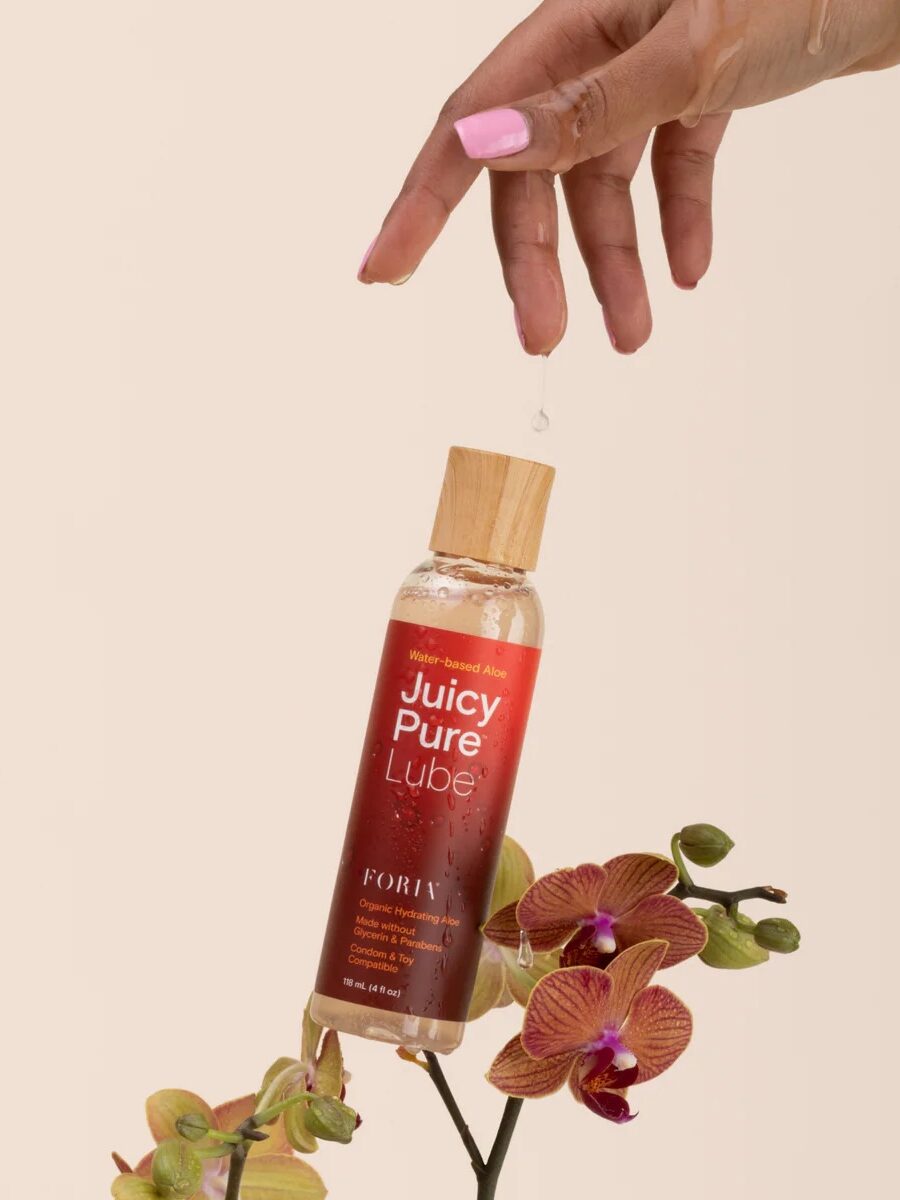 A hand with pink nails lets a drop of liquid fall onto a bottle of Juicy Pure Lube, which is balanced among purple orchids against a neutral background.