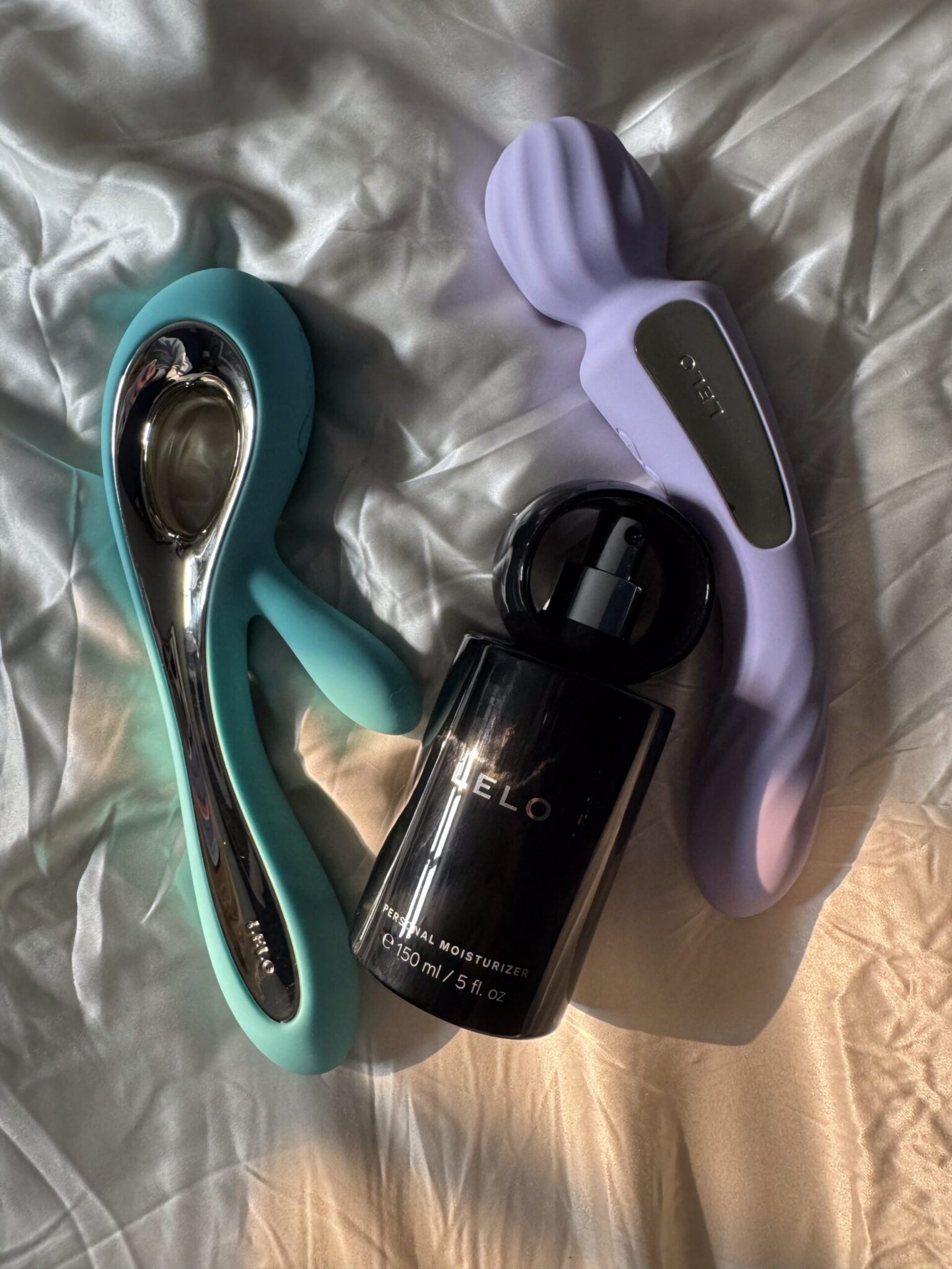 Two silicone vibrators and a black bottle of personal moisturizer on a white satin sheet, in natural sunlight.