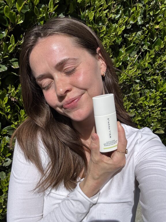 A woman with brown hair holds a white skincare bottle labeled "EVTILORSE" while standing outside in sunlight, smiling with her eyes closed. Green leafy bushes are in the background.