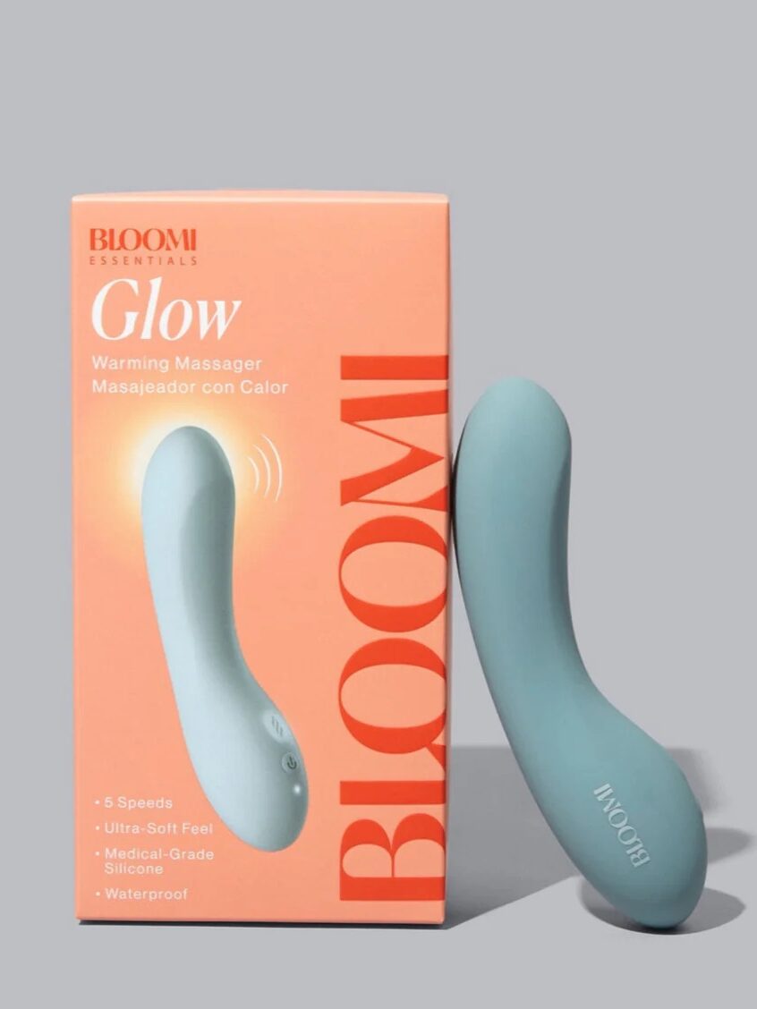 A Bloom Glow warming massager in light blue is shown next to its orange box packaging, which highlights features like 5 speeds and medical-grade silicone.