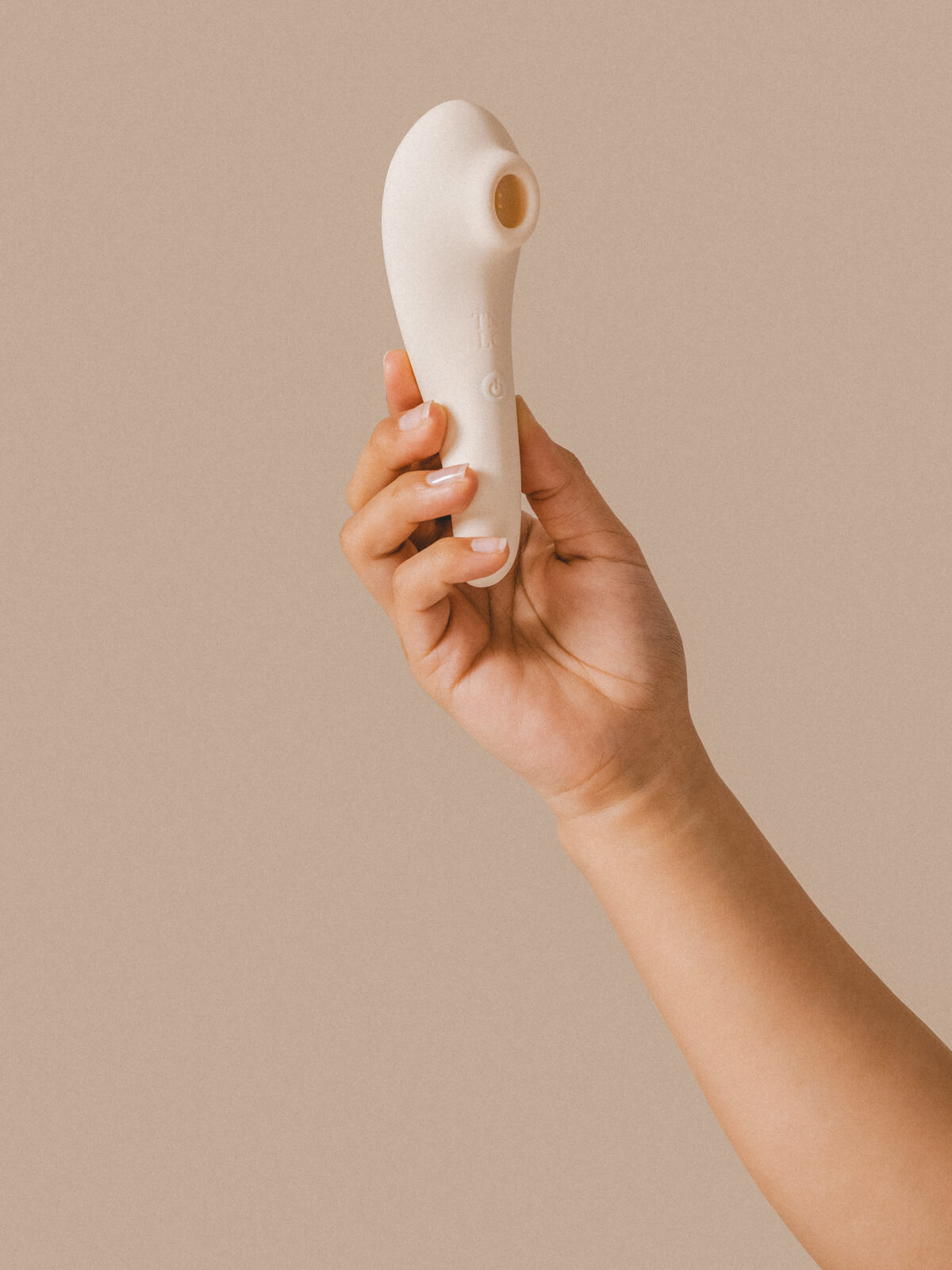 A hand holding a white, modern silicone personal massager with a rounded tip and a circular opening, set against a neutral beige background.