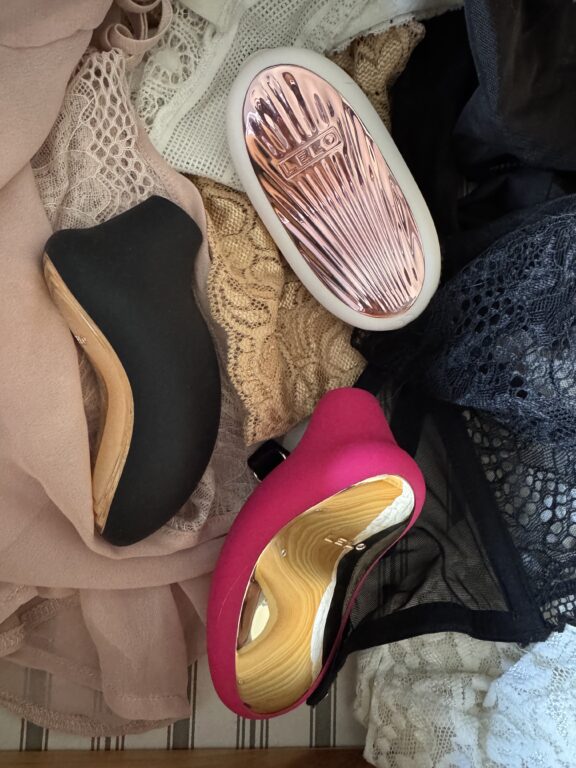 Three vibrators in black, pink, and rose gold rest on top of assorted lace and mesh fabrics in various colors.