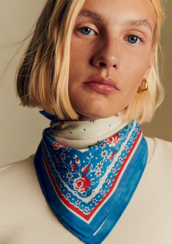 A woman with blonde hair wears a gold hoop earring and a blue and red floral scarf around her neck, over a cream-colored top, looking directly at the camera.