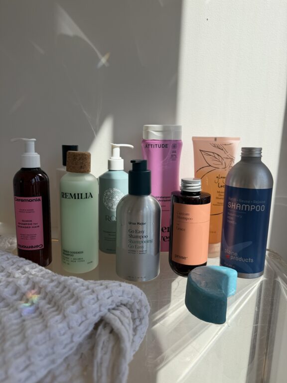 Various hair care products in colorful bottles and bars arranged on a clear surface with a folded white towel, lit by sunlight from the right.