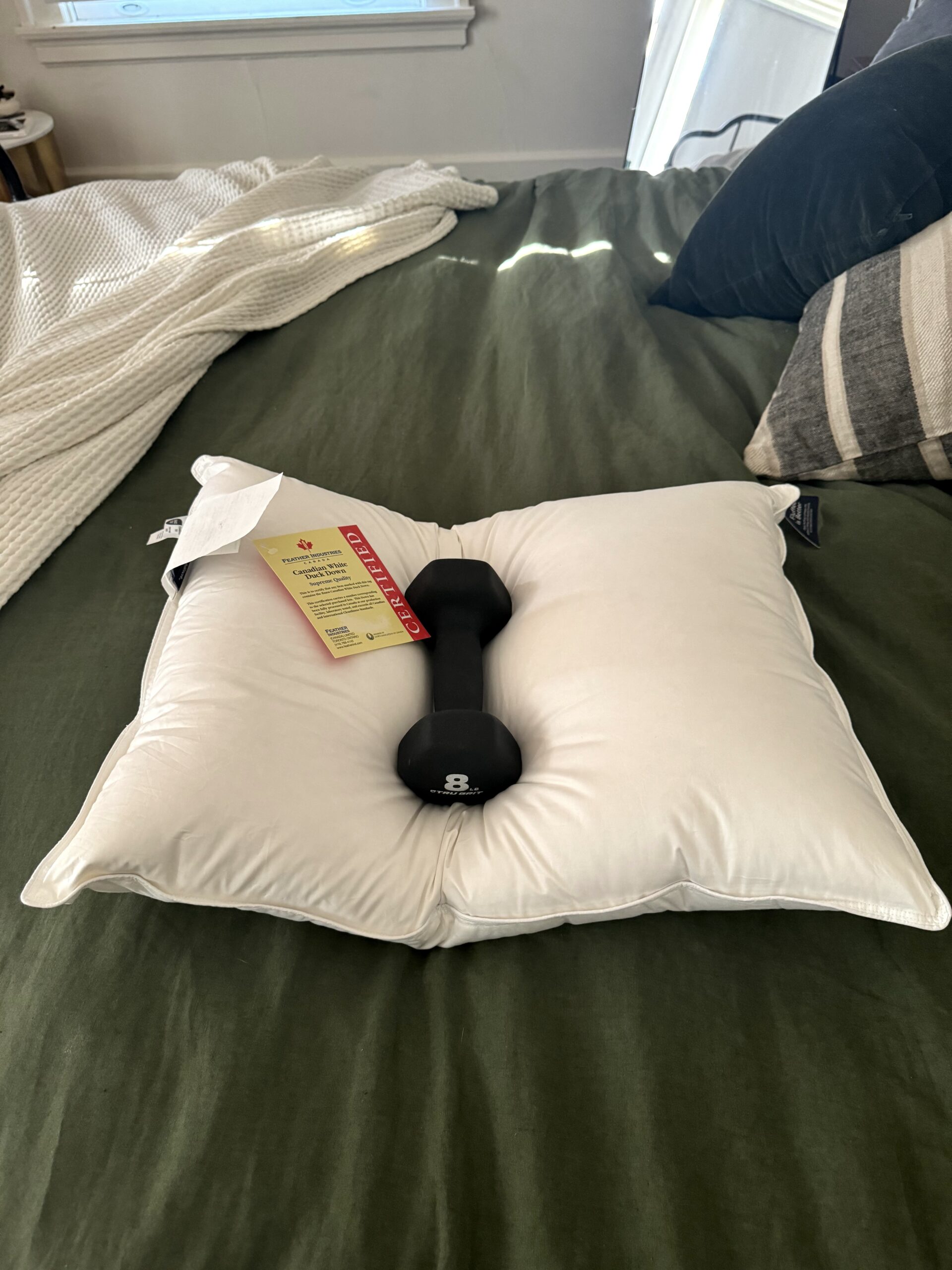 A white pillow with tags is placed on a green bedspread. An 8-pound black dumbbell sits on top of the pillow. A window and part of the bed are visible in the background.