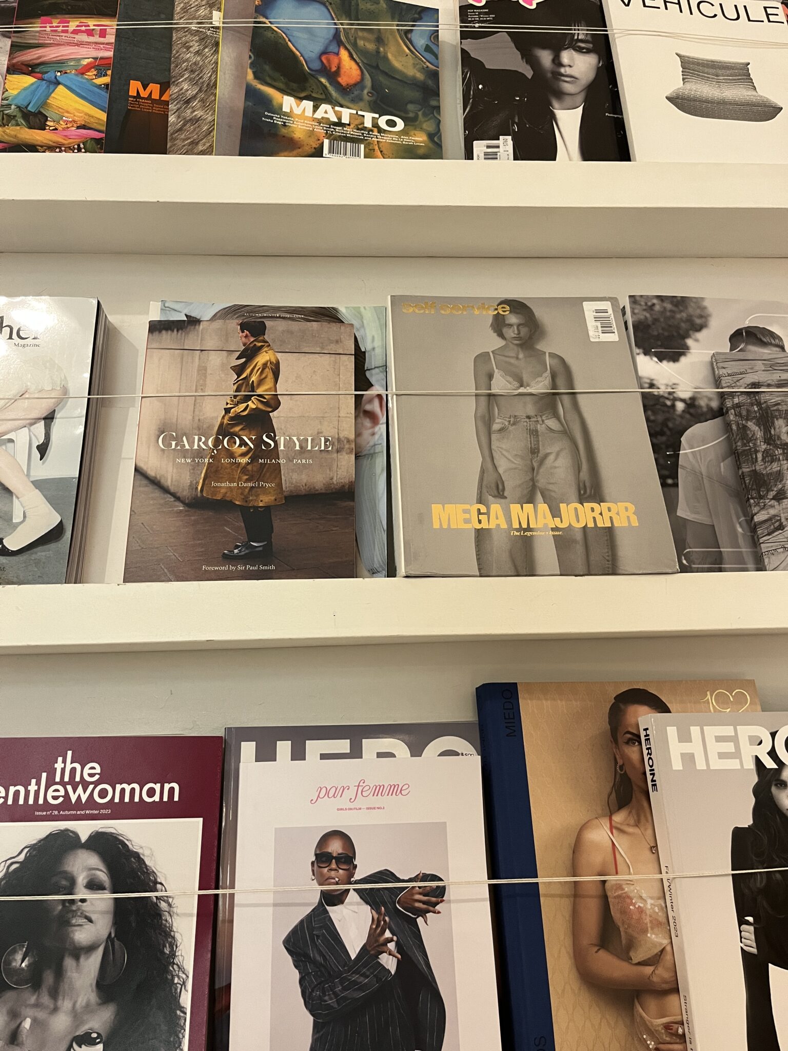 Magazines displayed on shelves, featuring various covers with models and fashion themes, including titles like "GARÇON STYLE," "MEGA MAJORRR," "the Gentlewoman," and "HERO.