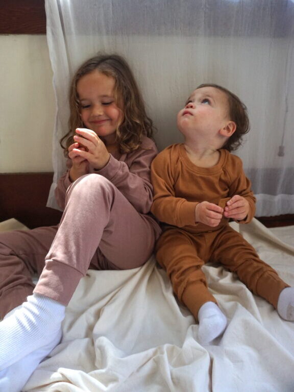 Two young children in pajamas sit on a blanket indoors; one child is smiling at an object in their hands, while the other looks up toward the window.