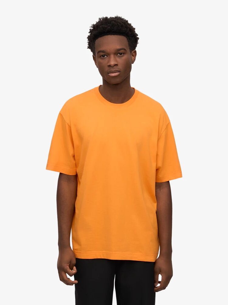 A person stands facing forward, wearing a plain orange T-shirt and black pants against a plain white background.