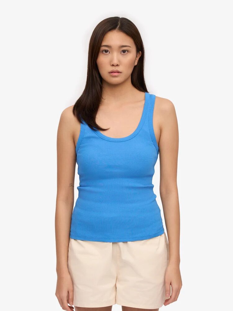 A woman with straight dark hair wears a blue tank top and beige shorts, standing against a plain white background.