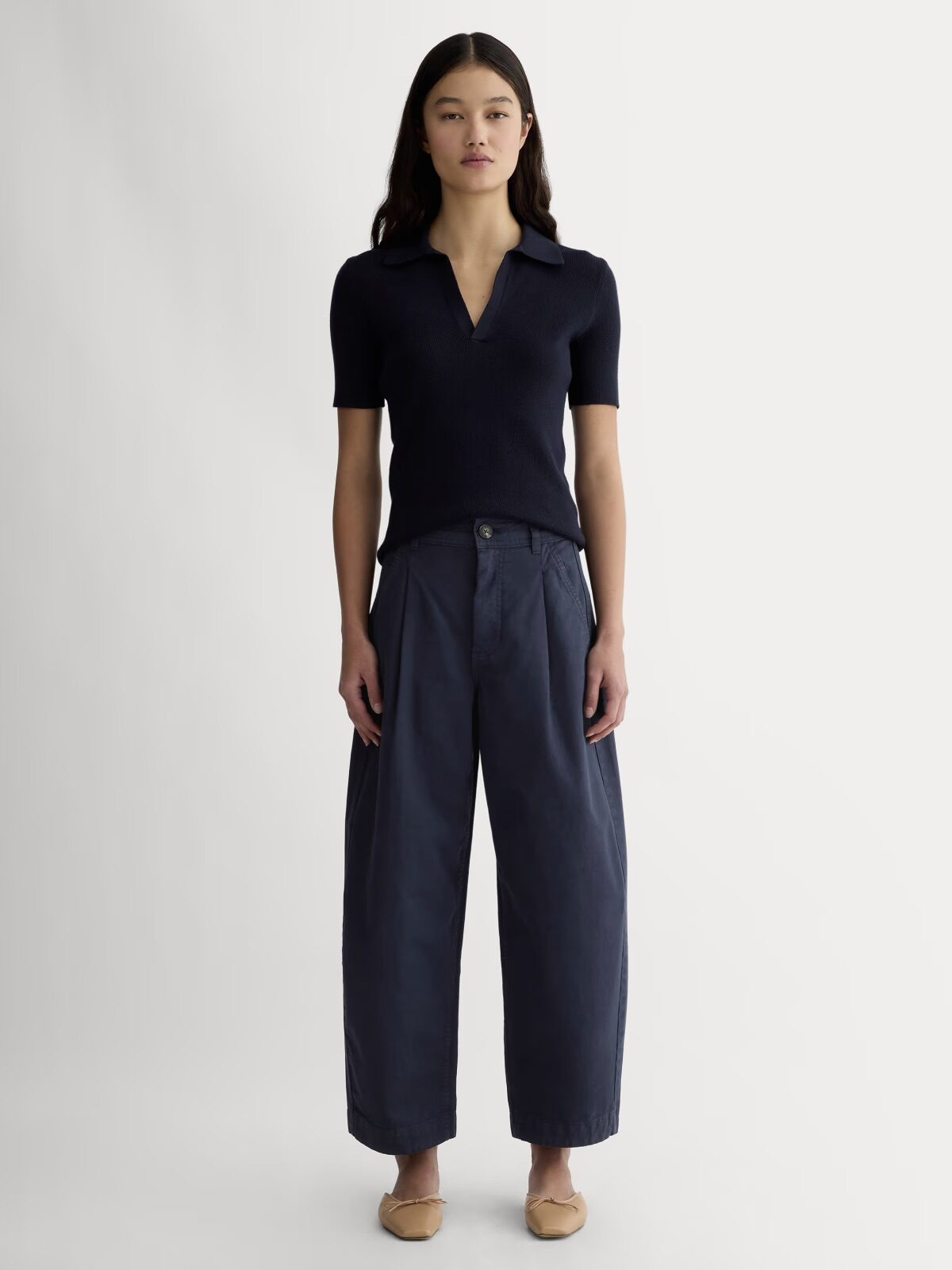 A woman stands facing forward, wearing a short-sleeve navy knit polo shirt, wide-leg navy pants, and beige flats, against a plain light background.