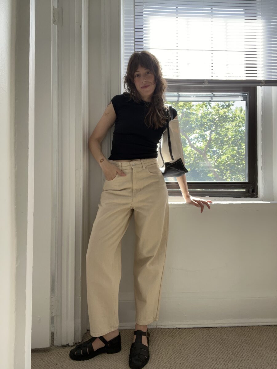 A woman in a black top and beige pants stands by a sunlit window with one hand resting on the windowsill.