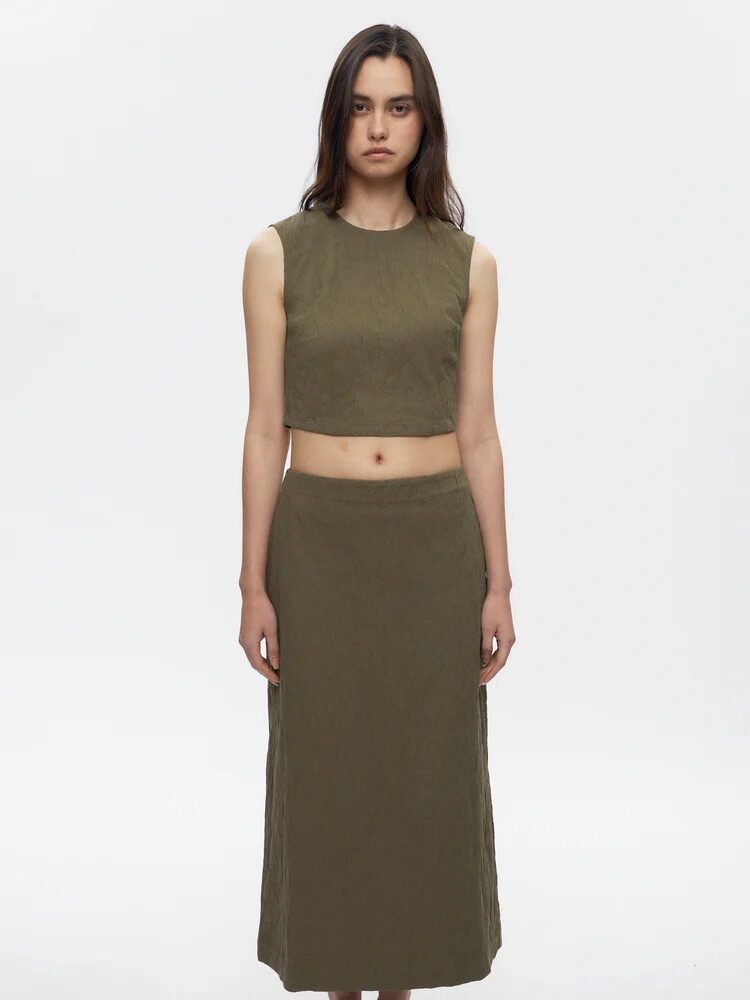 A woman stands against a plain background wearing an olive green sleeveless crop top and matching long skirt with beige flat shoes.